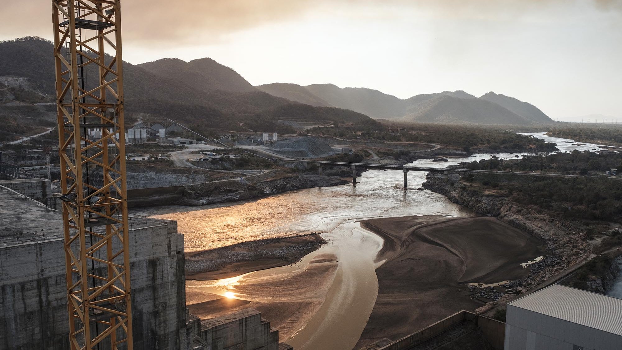 L'image montre un paysage industriel au bord d'une rivière. On y voit des collines et des montagnes en arrière-plan, enveloppées d'une légère brume. La rivière, au milieu, reflète la lumière du soleil, créant des scintillements sur sa surface. À gauche, il y a des structures en béton, probablement des installations industrielles. Un pont traverse la rivière, reliant des rives opposées. L'atmosphère semble calme et légèrement mélancolique en raison de la lumière dorée du coucher de soleil.