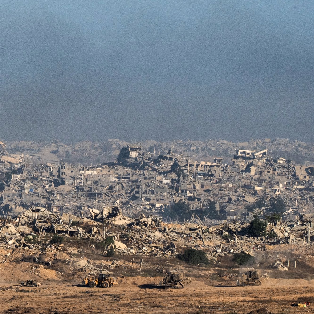 منظره‌ای از ویرانی وسیع، ساختمان‌های تخریب شده و دود در آسمان، با ماشین‌آلات ساختمانی در حال کار.