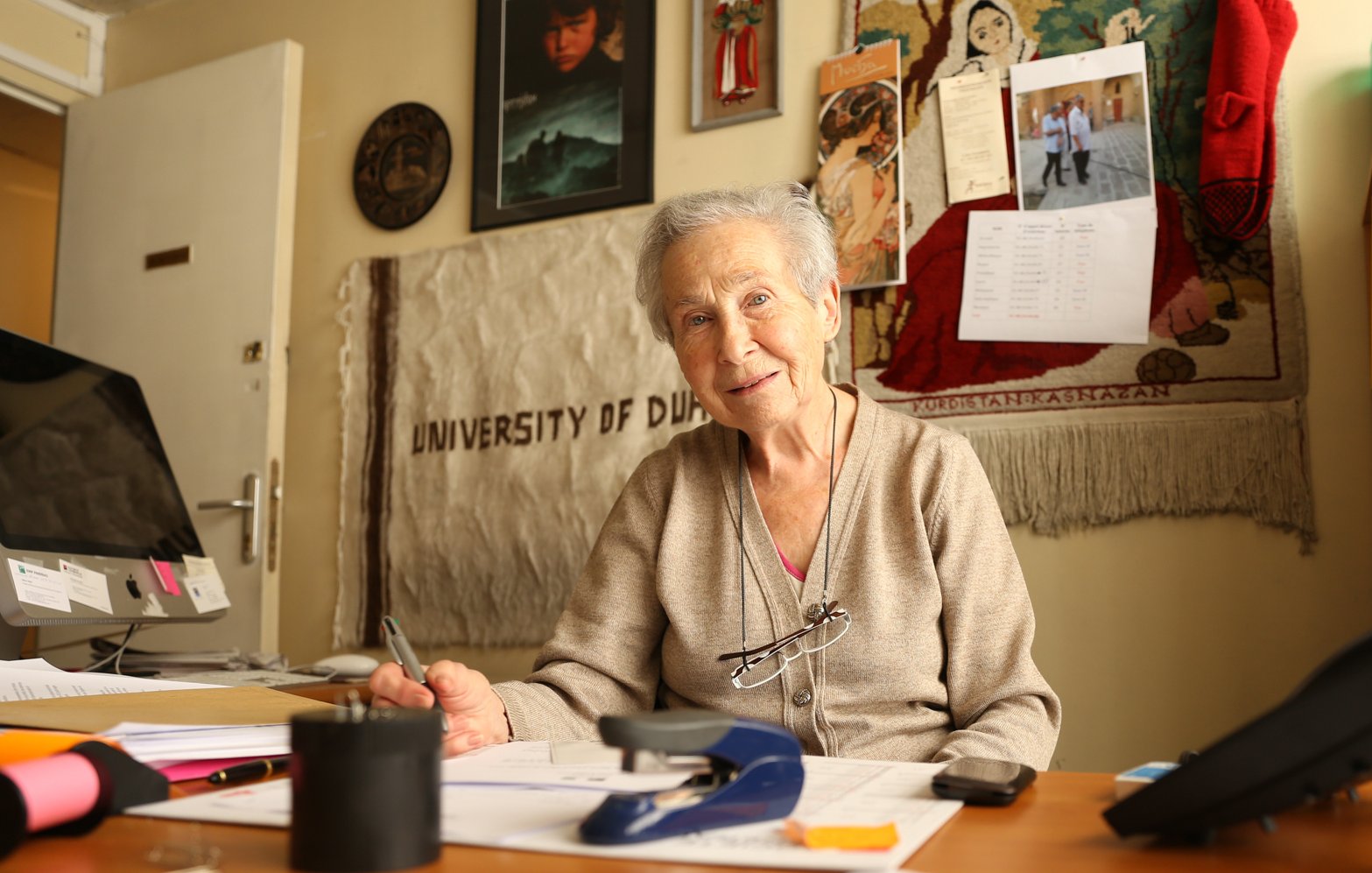 L'image montre une femme âgée assise à un bureau. Elle porte un pull beige et a des lunettes qui reposent sur son nez. Son sourire est chaleureux et accueillant. Le bureau est en désordre avec des papiers éparpillés, un agrafeuse et un verre. En arrière-plan, on aperçoit divers objets accrochés au mur, y compris un calendrier et des photos. On peut aussi distinguer une toile ou un tissu qui mentionne "UNIVERSITY OF DUBAI". L'atmosphère est conviviale et studieuse.