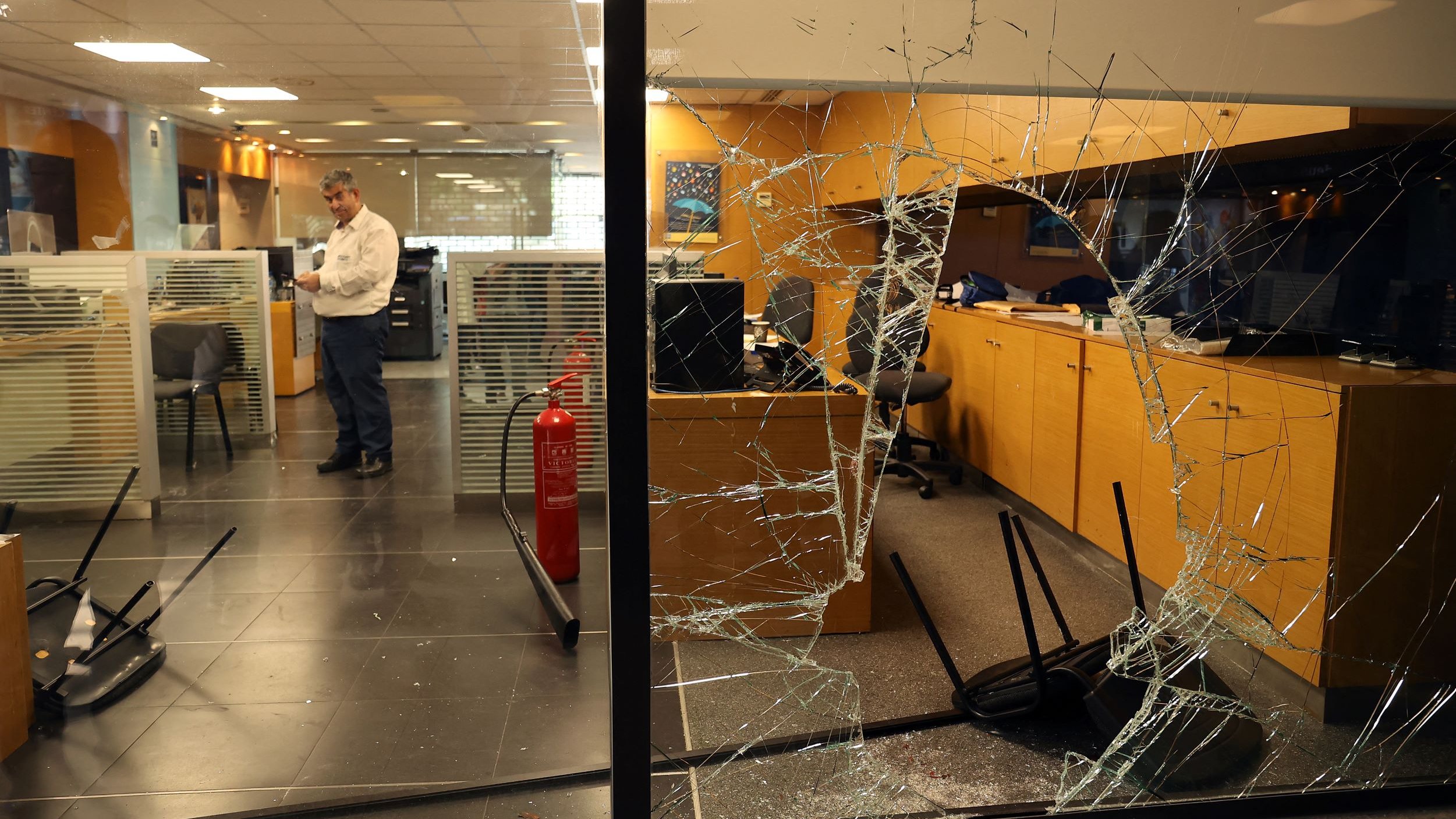 تظهر الصورة داخل مكتب يتضمن مشاهد من الفوضى، حيث يوجد زجاج مكسور على أحد النوافذ، مما يشير إلى وجود حادث أو تخريب. في الخلفية، يمكن رؤية أثاث مكتبي، مثل الطاولات والكراسي، وبعضها مقلوب. يوجد أيضًا شخص واحد يرتدي قميصًا فاتح اللون، يبدو وكأنه يتفقد الوضع داخل المكتب. الجو العام يوحي بحالة من الاضطراب أو الهجوم.