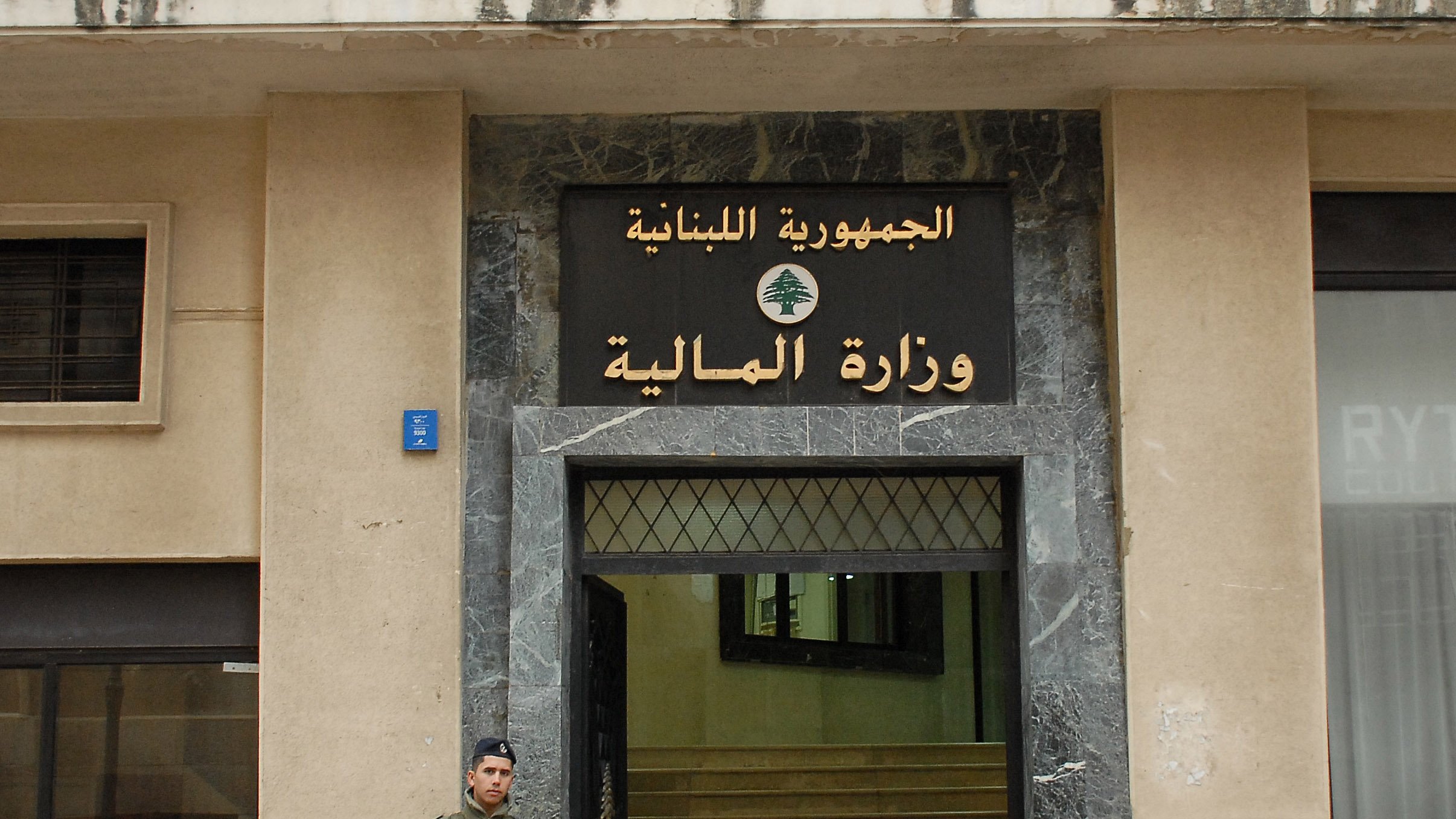The image shows a soldier standing outside a building with a large sign displaying the words "الجمهورية اللبنانية" (Lebanese Republic) and "وزارة المالية" (Ministry of Finance) in Arabic. The building has a modern façade with a marble entrance. The soldier is wearing military uniform and holding a weapon, indicating a security presence at the site. The overall setting suggests a government location in Lebanon.