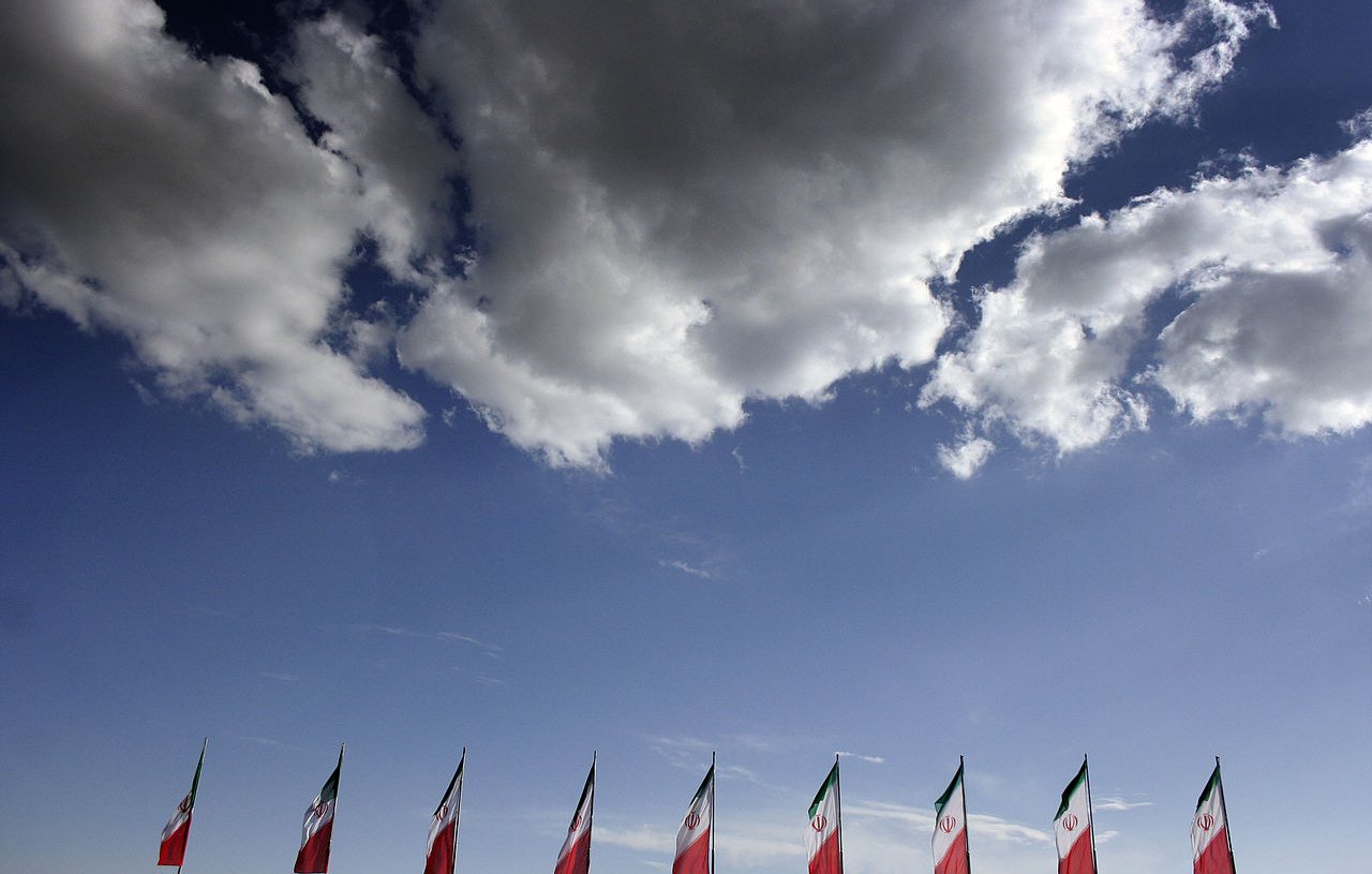 The image features a clear blue sky with fluffy white clouds. In the foreground, several flags, likely representing Mexico, are lined up. The flags prominently display the colors of the Mexican flag, which includes green, white, and red, along with the national emblem. The overall scene conveys a sense of patriotism against a serene backdrop.