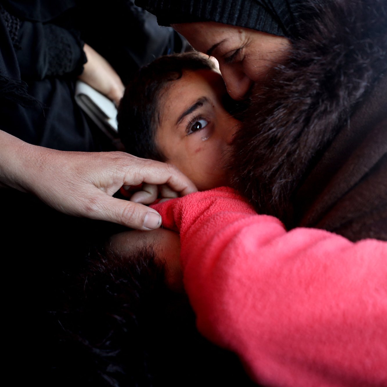 L'image montre un moment touchant entre un enfant et un adulte. L'adulte semble embrasser l'enfant, qui a l'air attentionné et gaze directement vers l'objectif. On peut aussi voir quelques mains, une d'elles touchant l'épaule de l'enfant. L'ambiance est empreinte d'émotion et de tendresse. Les vêtements des figures suggèrent un environnement austère.