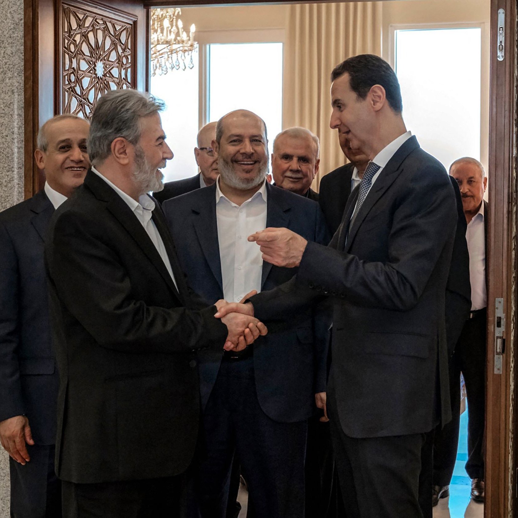 L'immagine mostra un gruppo di uomini in un contesto formale, probabilmente in un ufficio o in un luogo ufficiale. Al centro, due uomini si stringono la mano, mentre gli altri li osservano con espressioni di interesse o approvazione. Gli abiti sono eleganti e i colori sono sobri, creando un'atmosfera seria e professionale. Le pareti e l'arredamento suggeriscono un ambiente di alto livello, con dettagli architettonici visibili sullo sfondo.