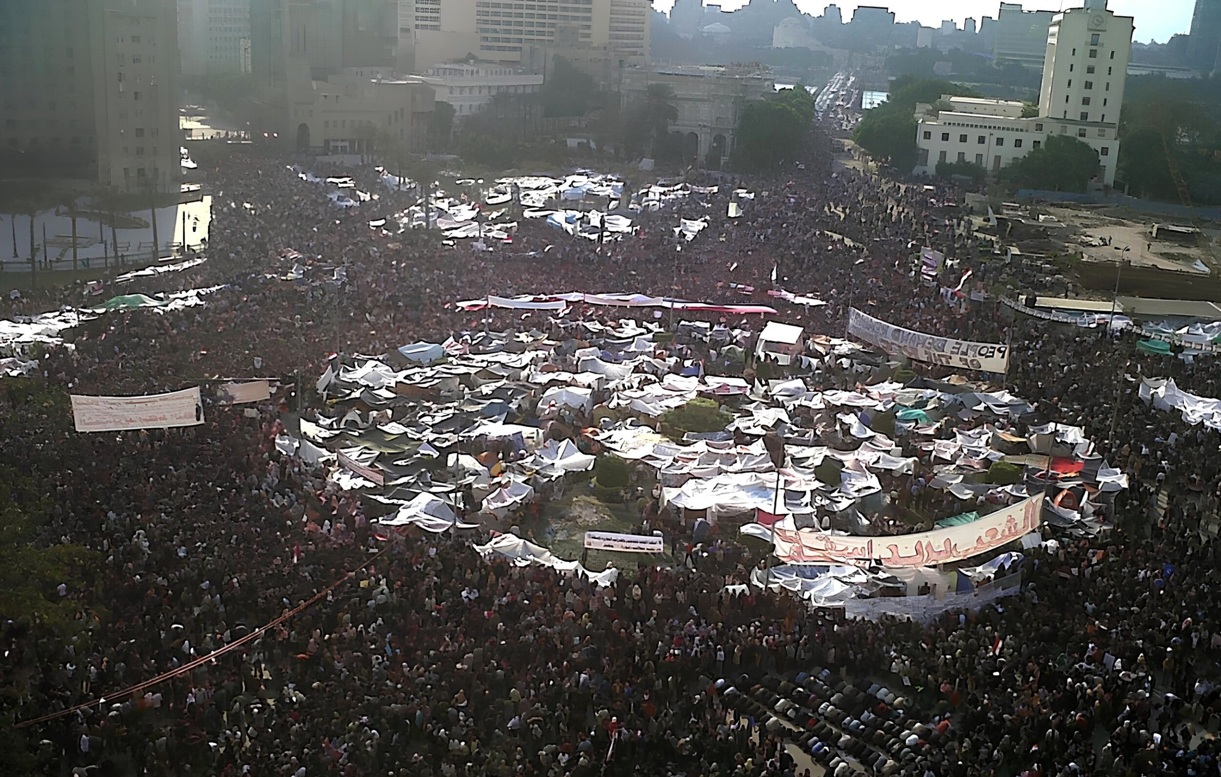 Una grande folla si raduna in una piazza, con tende e striscioni.
