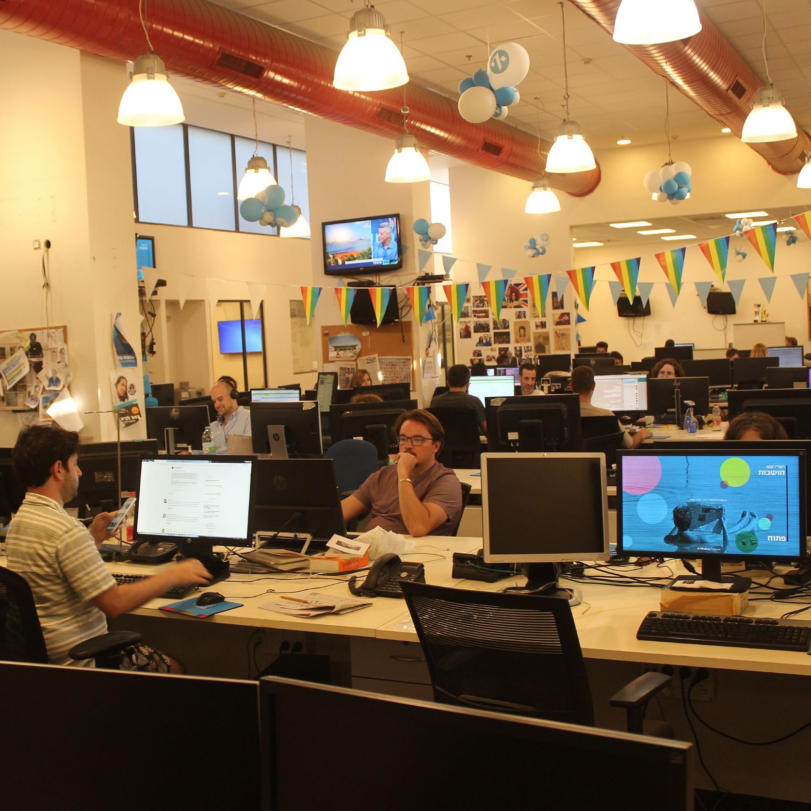 La imagen muestra una oficina moderna y espaciosa. Hay varias estaciones de trabajo con computadoras y pantallas en escritorios. Algunas personas están concentradas en sus tareas mientras que otras parecen colaborar o comunicarse. El ambiente es iluminado y decorado con globos y banderas, lo que sugiere que podría ser un lugar festivo o creativo. Las paredes tienen fotografías y otros elementos decorativos, añadiendo un toque personal al espacio laboral.