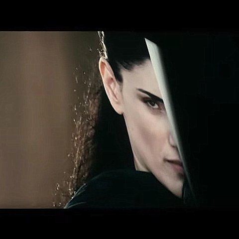 L'image montre une femme avec une expression intense et déterminée. Elle est à moitié cachée derrière un objet, ce qui accentue son regard perçant. Ses cheveux sont longs et foncés, et son attitude suggère qu'elle est dans une situation de confrontation ou de défense. L'éclairage met en avant son visage et crée une ambiance mystérieuse.