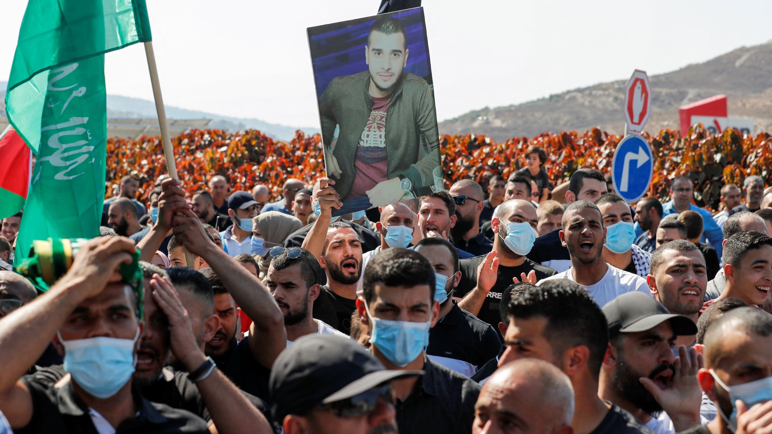 The image depicts a large crowd participating in a protest or demonstration. Many individuals are holding up flags, including green ones, and a prominent figure is holding a portrait of a man. The crowd appears to be engaged and unified, with some people wearing face masks. The background features hills, adding to the overall atmosphere of the event.