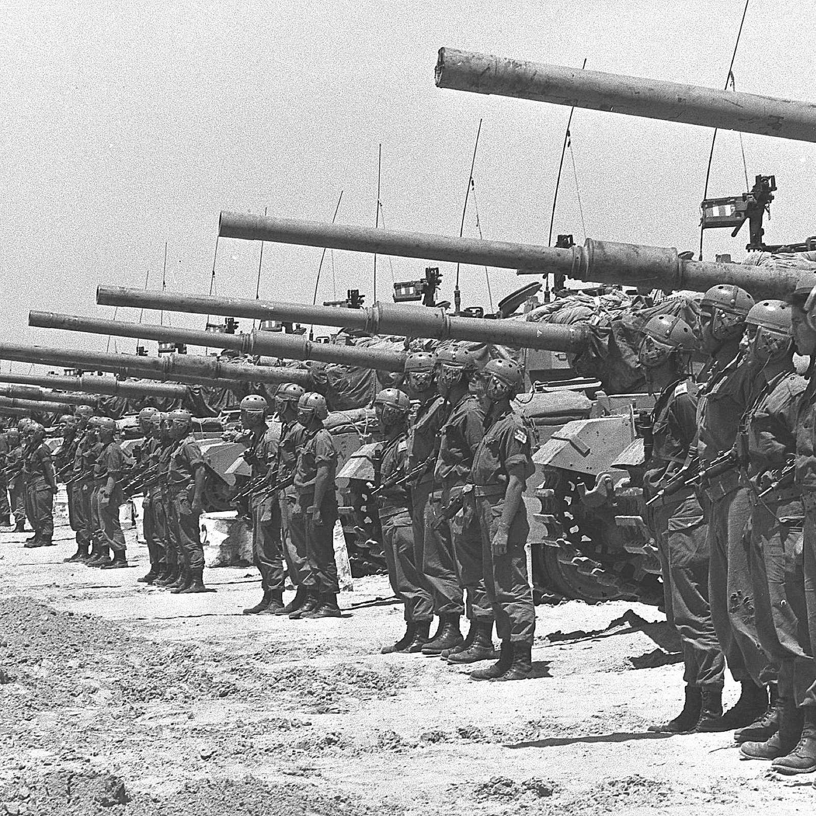 L'image montre un groupe de soldats alignés de manière formelle, portant des uniformes militaires et des casques. Ils sont positionnés devant une rangée de chars d'assaut, qui sont également alignés. Le paysage semble désertique, sous un ciel clair. L'atmosphère évoque une préparation militaire, peut-être pour une parade ou une démonstration de force. Les chars sont équipés de canons pointés vers l'avant, ce qui ajoute à l'impression de puissance et de militarisation de la scène.