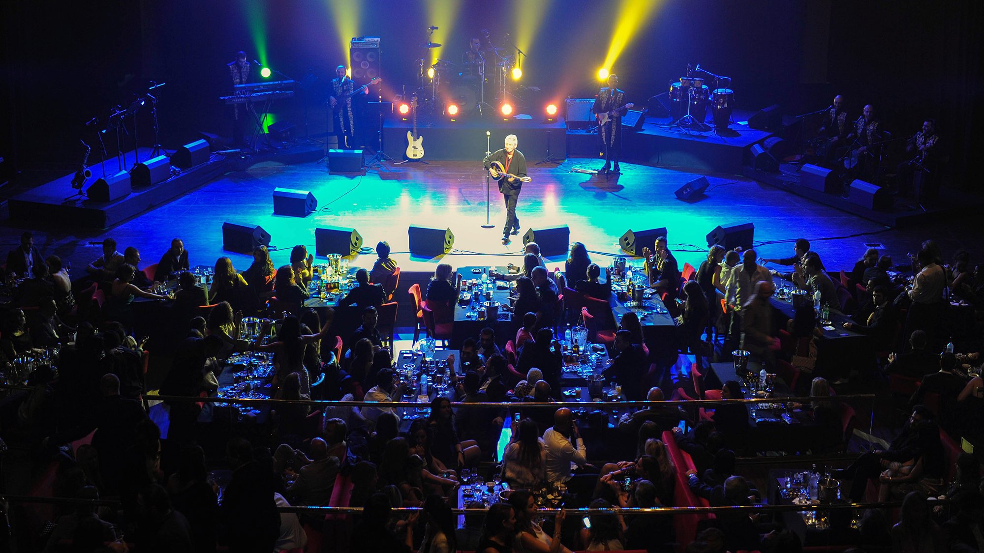 L'image montre une scène de concert animée. Au premier plan, un artiste se produit sur scène, entouré de lumières colorées et de spots éclairant l'espace. En arrière-plan, on peut voir des musiciens qui l'accompagnent. La salle est pleine de spectateurs assis à des tables, savourant leur soirée avec des repas et des boissons. L'ambiance est festive et chaleureuse, avec des gens qui semblent apprécier la performance.