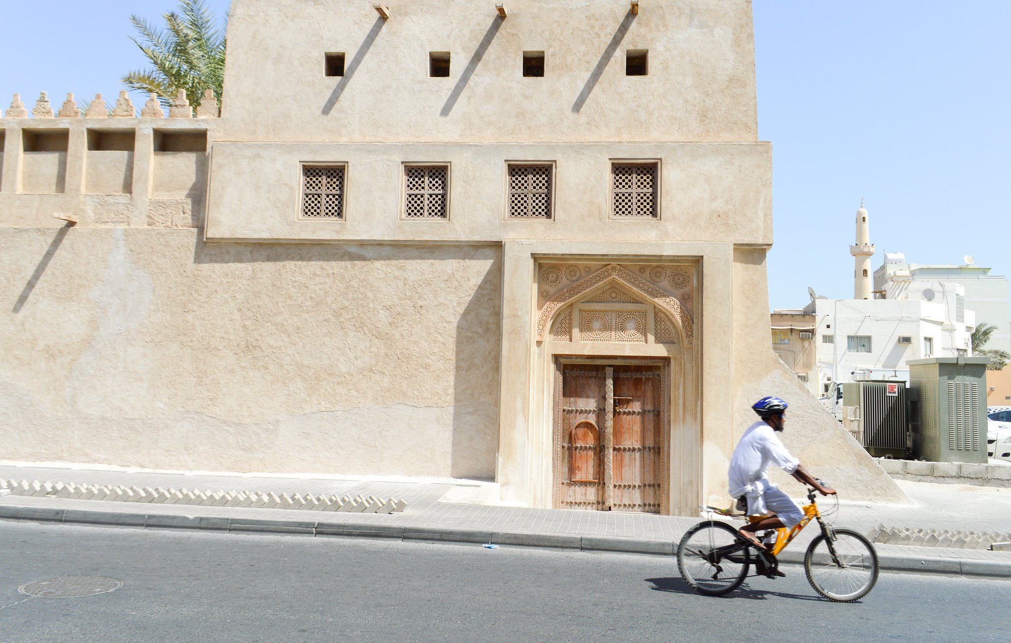 L'image montre un homme à vélo qui passe à côté d'un bâtiment au style architectural traditionnel. Ce bâtiment, de couleur sable, présente des murs texturés et des fenêtres ornées de motifs. On peut apercevoir une grande porte en bois sculpté, typique de l'architecture ancienne. En arrière-plan, on voit une mosquée avec un minaret, ce qui évoque une ambiance tranquille et culturelle dans une ville du Moyen-Orient. Le ciel est dégagé et ensoleillé.