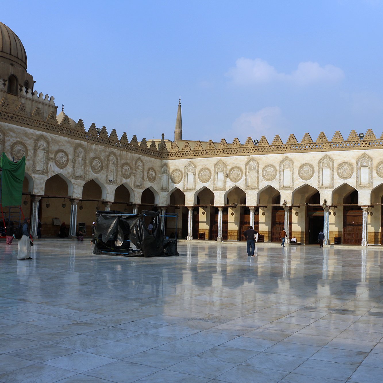 L'image montre une cour spacieuse d'un bâtiment historique, probablement un lieu de culte ou une mosquée. Le sol est en marbre poli, reflétant la lumière du ciel. On peut observer des arches élégantes et de nombreuses colonnes tout autour, ornées de motifs architecturaux détaillés. Au fond, on aperçoit un dôme et des minarets qui s'élèvent dans le ciel. La scène est calme, avec quelques personnes se déplaçant dans l'espace. Le ciel est clair, parsemé de nuages légers.