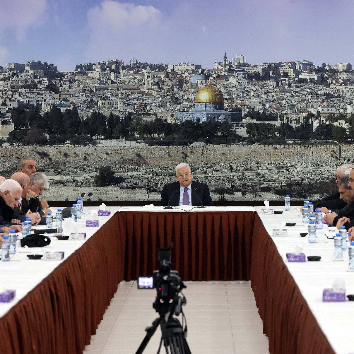 تظهر الصورة مجموعة من الأشخاص يجلسون حول طاولة كبيرة في قاعة اجتماع. في خلفية الصورة، يُمكن رؤية منظر لمدينة القدس مع قبة الصخرة. يبدو أن الجلسة تتعلق بقضية هامة، حيث يركز الجميع على شخص يجلس في المنتصف. الطاولة مرتبة بشكل مرتب، مع وجود زجاجات مياه وأكواب على الطاولة. يتمحور اللقاء حول موضوع قد يكون له تأثير كبير على المجتمع أو السياسة.