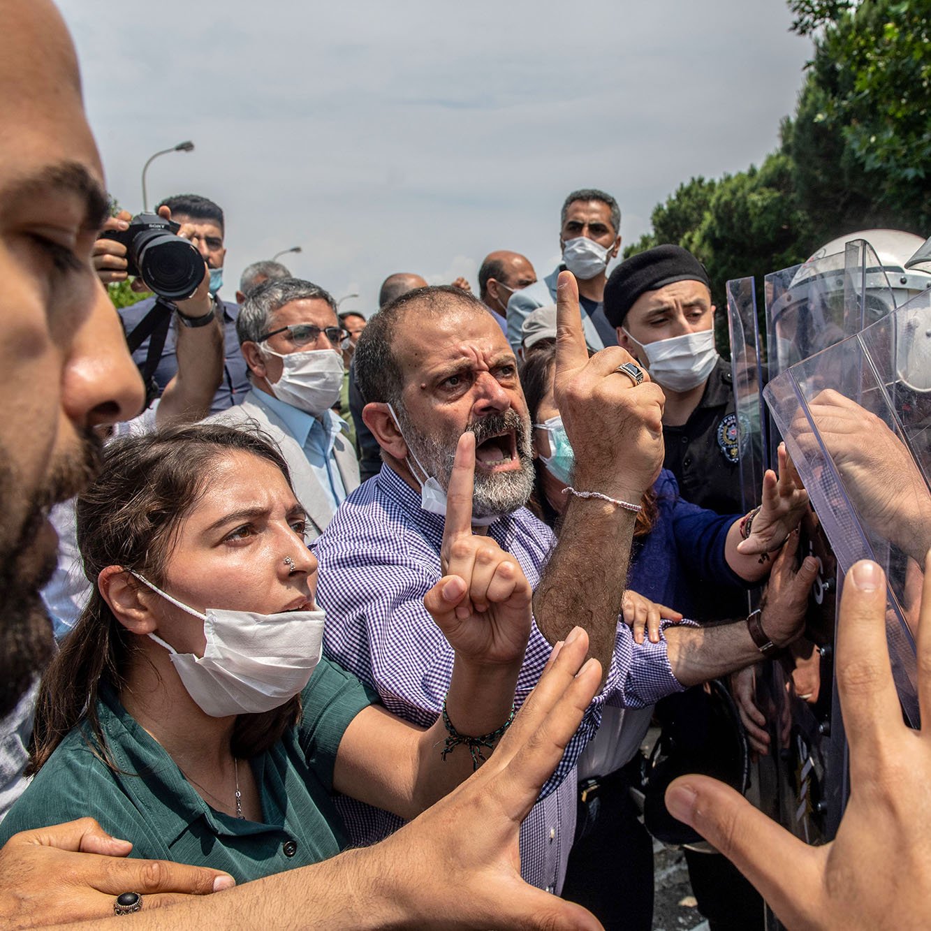L'image montre une scène de tension entre des manifestants et des forces de l'ordre. Au premier plan, plusieurs personnes expriment leur désaccord, avec des gestes vifs et des expressions de colère ou de frustration. Certaines personnes portent des masques, probablement en raison de préoccupations sanitaires. En arrière-plan, des membres des forces de l'ordre, équipés de casques et de boucliers, semblent faire face aux manifestants. L'environnement est urbain, et le ciel est partiellement nuageux. L'atmosphère est chargée d'émotion et de conflit.