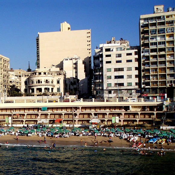 L'image montre une plage animée avec de nombreux baigneurs profitant du soleil et de la mer. On peut voir des parasols disposés sur le sable, ainsi que des hôtels et des immeubles en arrière-plan. L'architecture des bâtiments est variée, avec des structures anciennes côtoyant des constructions plus modernes. Le ciel est dégagé, et l'atmosphère est vivante, typique d'une destination balnéaire populaire.
