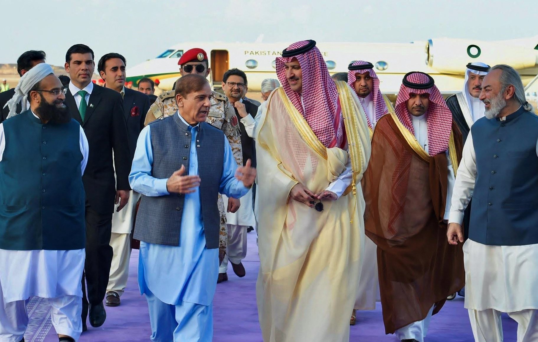 L'image montre un groupe de personnes en tenue formelle, marchant ensemble sur un tapis violet. On peut voir des hommes portant des costumes traditionnels et des vêtements d'apparat, avec certains en toges ou en robes. En arrière-plan, un avion privé est stationné, indiquant un contexte officiel ou diplomatique. L'atmosphère semble conviviale, avec les participants engagés dans une conversation.