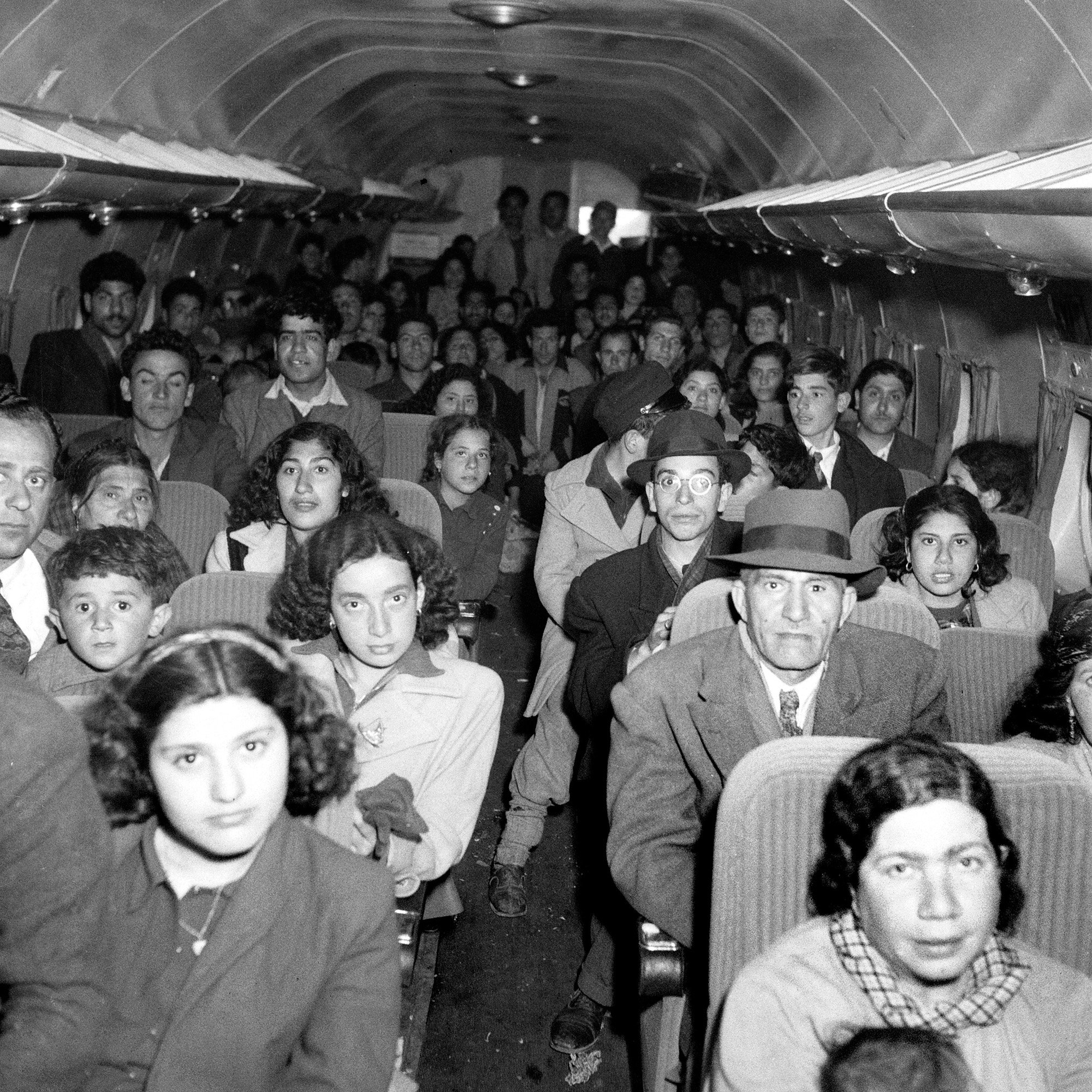 L'image montre l'intérieur d'un avion rempli de passagers. On peut voir un groupe diversifié de personnes, y compris des adultes et des enfants, assis sur des rangées de sièges. Les expressions des passagers varient, certains semblent sérieux, tandis que d'autres regardent droit devant. L'ambiance semble être celle d'un voyage, probablement dans un contexte historique. L'éclairage de l'image est en noir et blanc, ce qui lui donne une atmosphère vintage.