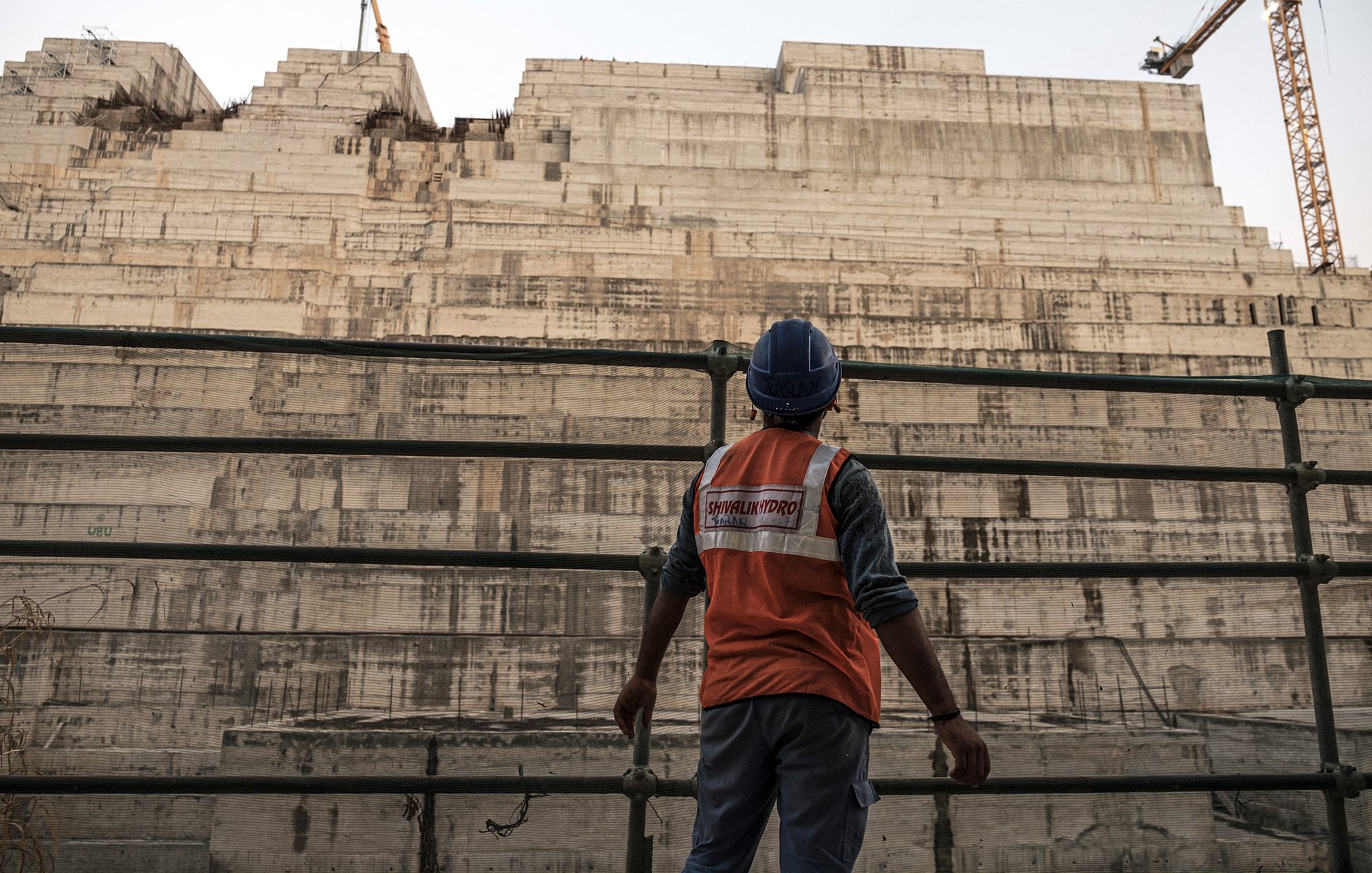 تظهر في الصورة عامل بناء يرتدي سترة واقية زاهية اللون وخوذة، وهو يقف أمام هيكل كبير من الخرسانة يبدو أنه في مرحلة الإنشاء. خلفه، تُلاحظ طبقات من المواد البنائية، مما يشير إلى مشروع بناء ضخم. يظهر في الخلفية معدات البناء مثل رافعة، مما يضيف إلى جو العمل المكثف والحركة في المكان. السماء تميل إلى اللون الأزرق، مما يوحي بأن الوقت هو إما الصباح الباكر أو بعد الظهر.