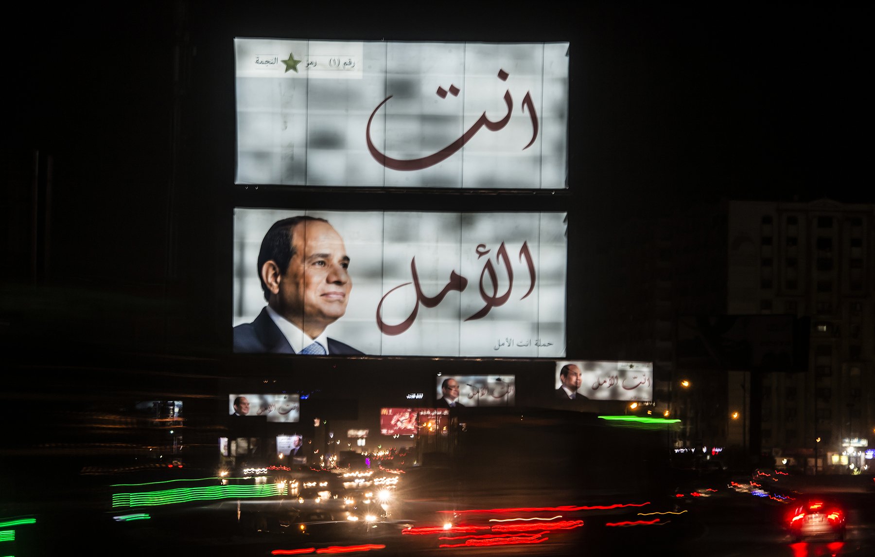 L'image montre un grand panneau d'affichage lumineux à la nuit tombante. Sur ce panneau, on peut voir un portrait d'un homme en costume. Les mots arabes "أنت الأمل" et "أنت" sont écrits en grandes lettres, indiquant un message fort. En bas, il y a une autre image de l'homme. L'environnement est animé, avec des lumières de voitures qui passent en bas, créant un effet de mouvement et une ambiance urbaine nocturne.
