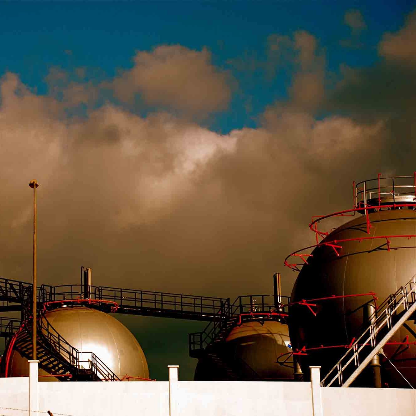 این تصویر نمایی از یک تأسیسات صنعتی را نشان می‌دهد که شامل چند مخزن بزرگ به شکل کروی است. مخازن روی سکویی قرار دارند و برخی از آن‌ها داکت‌ها و لوله‌های پیچیده‌ای دارند که به آن‌ها متصل شده‌اند. آسمان در پس‌زمینه کمی ابری است و رنگ‌ها به طور کلی گرم و متنوع به نظر می‌رسند. اجزای صنعت و بافت‌های مختلف تأسیسات به خوبی در تصویر دیده می‌شوند.