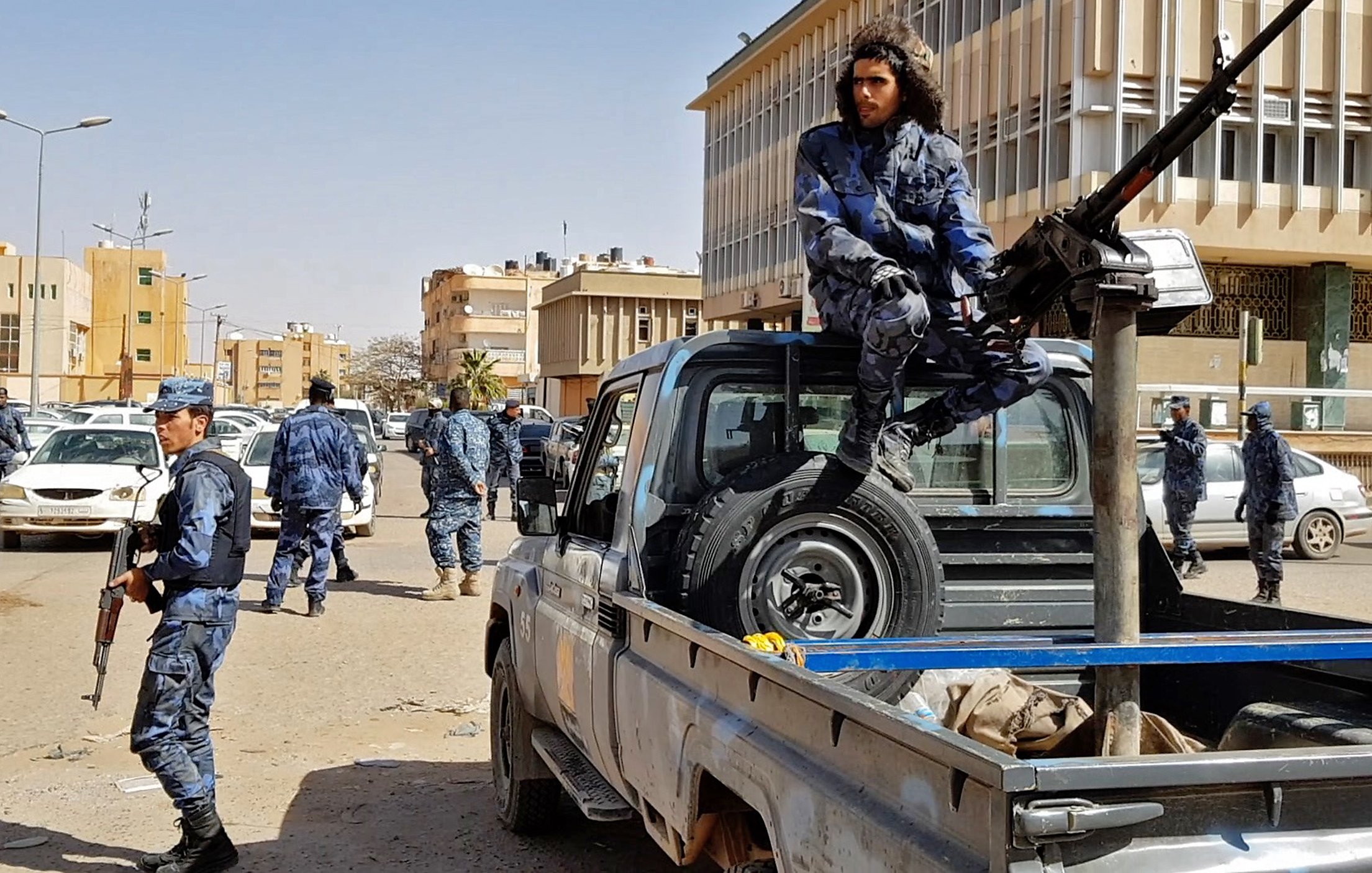 L'image montre une scène urbaine impliquant des forces de sécurité. On peut y voir plusieurs policiers en uniforme, certains armés, se déplaçant autour de véhicules. Un homme est assis sur le plateau d'une camionnette, près d'une arme lourde montée sur le véhicule. L'environnement semble être une ville moderne, avec des bâtiments en arrière-plan. L'ambiance générale de l'image suggère une opération de sécurité ou une situation de contrôle.
