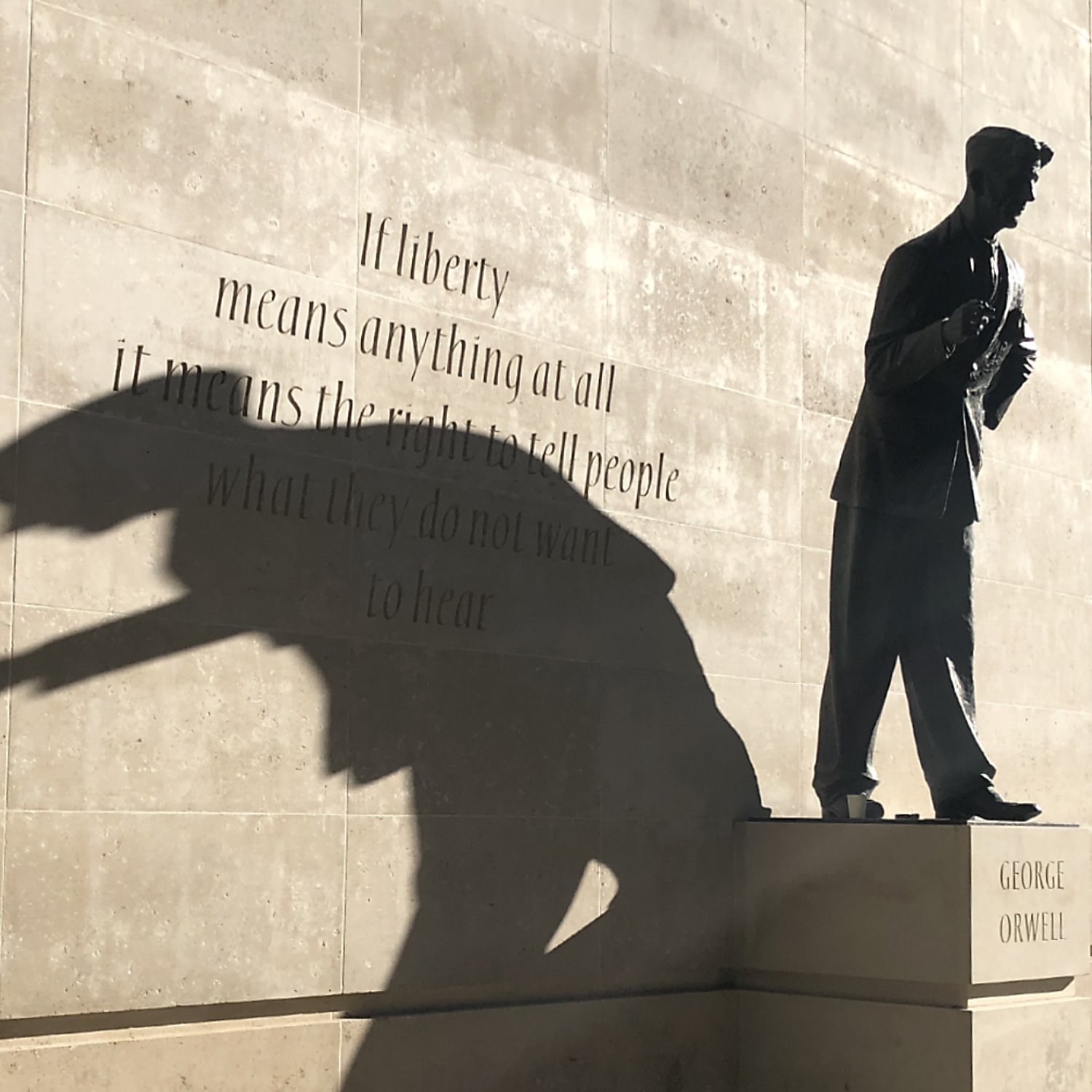 L'image montre une sculpture d'une silhouette d'un homme, probablement un écrivain, sur un socle avec le nom "GEORGE ORWELL". À côté de la statue, il y a une citation en anglais sur le thème de la liberté. La lumière crée une ombre qui semble projeter une forme dynamique, ajoutant une dimension artistique à la scène. Le mélange de texte et de sculpture évoque des réflexions sur la liberté d'expression.