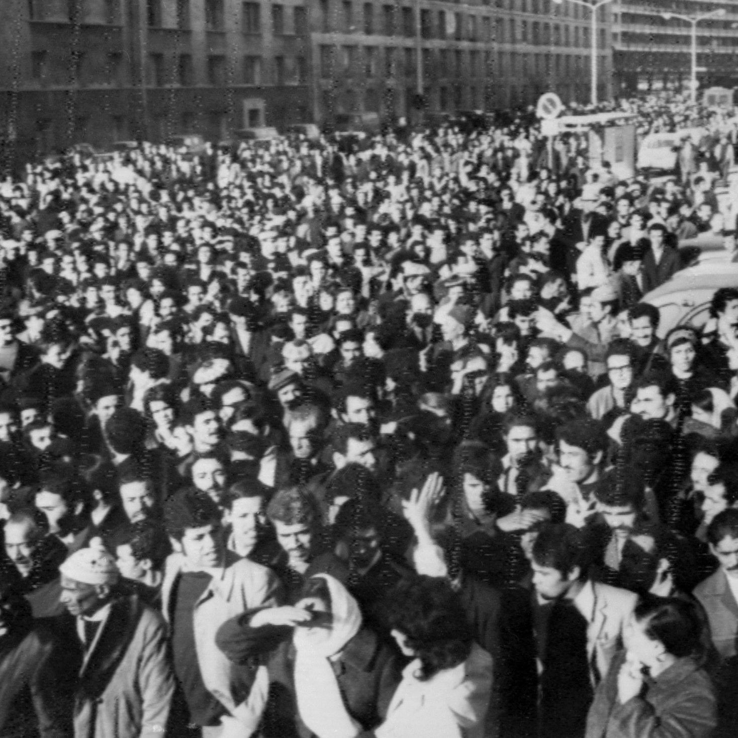 The image depicts a large crowd of people gathered in an urban setting, likely during a significant event or protest. The crowd appears to be densely packed, filling the street and surrounding areas. Many individuals are dressed in formal or semi-formal attire, indicating the seriousness of the occasion. The atmosphere seems charged, reflecting a sense of unity and purpose among the participants. Buildings line the background, and there are vehicles partially visible, suggesting a city environment. The black-and-white effect adds a historical or dramatic tone to the scene.