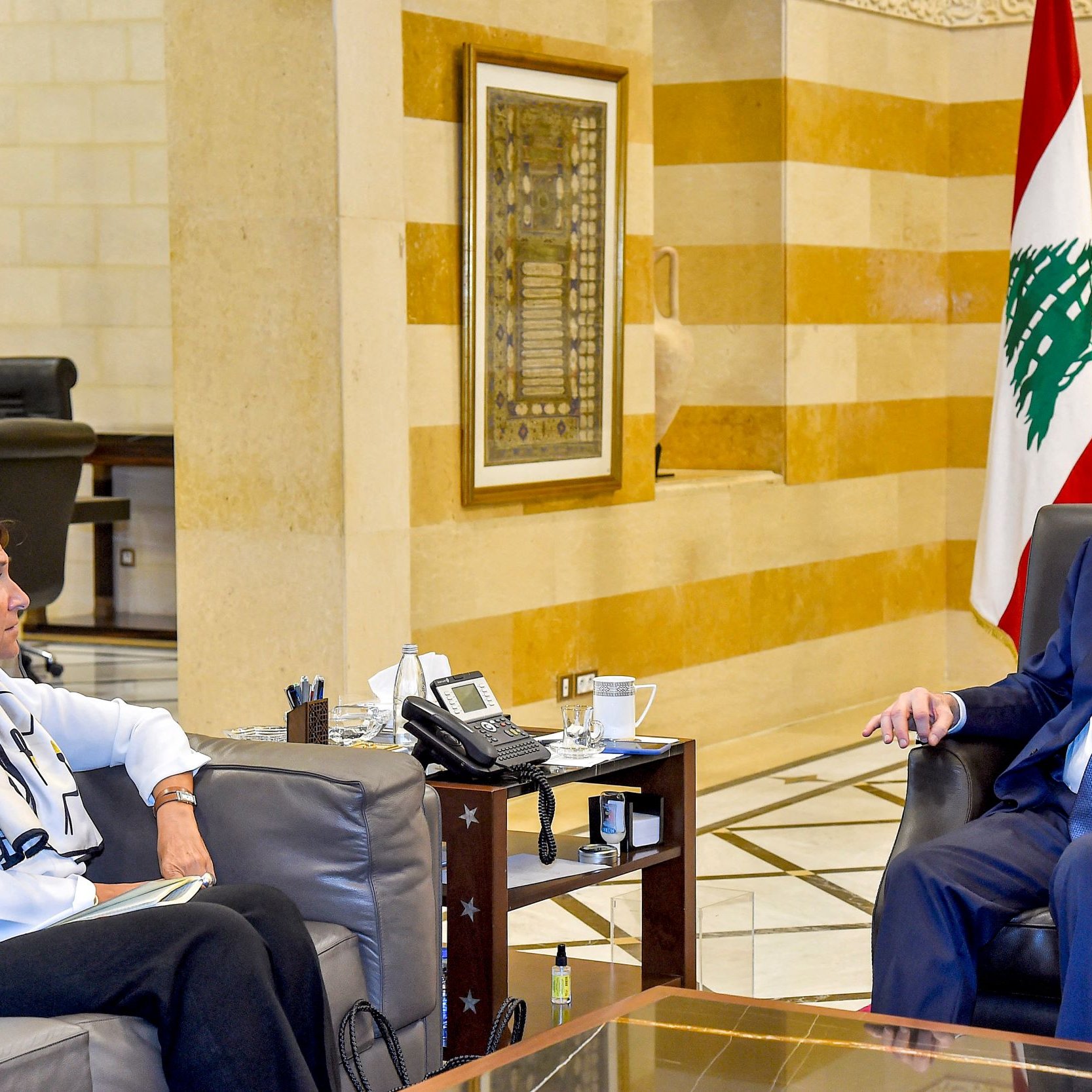 الصورة تظهر اجتماعًا داخل مكتب. يجلس رجل وامرأة على أريكتين. الرجل يبدو في منتصف العمر ويرتدي بدلة رسمية. المرأة ترتدي ملابس عادية، ويبدو أنها تشارك في الحوار. في الخلفية، تُرى ألوان العلم اللبناني، مما يدل على أن الاجتماع قد يكون ضمن إطار حكومي أو رسمي. الأثاث والتصميم الداخلي يعبّران عن الفخامة.