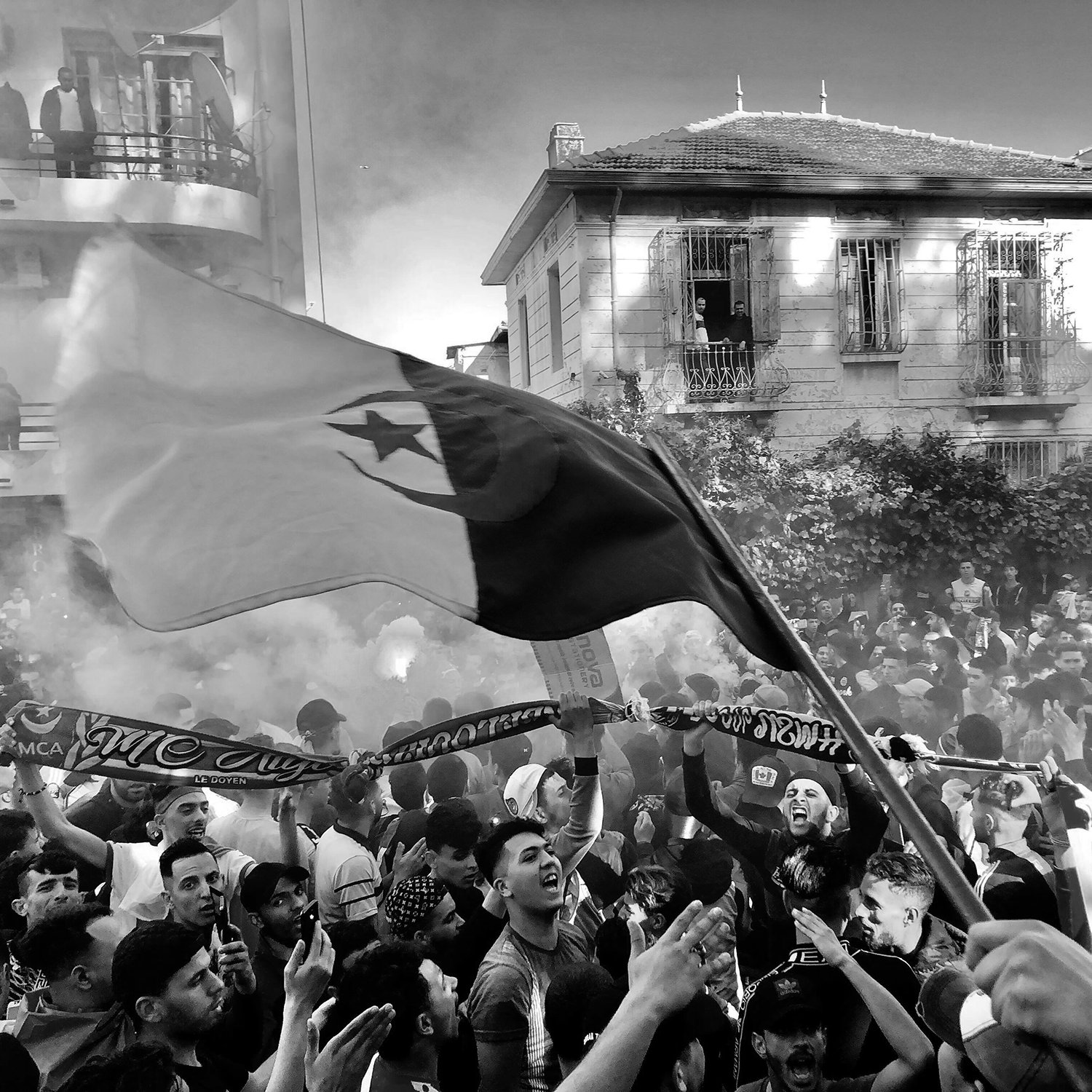 The image depicts a large crowd gathered in an urban setting, likely during a festive or protest event. People are seen waving flags and holding scarves, creating a vibrant atmosphere. Smoke is rising in the air, possibly from flares or fireworks, adding to the intensity of the scene. The buildings in the background have a mix of architectural styles, hinting at a lively neighborhood. The overall mood appears energetic and spirited, with individuals showing strong emotions and camaraderie.