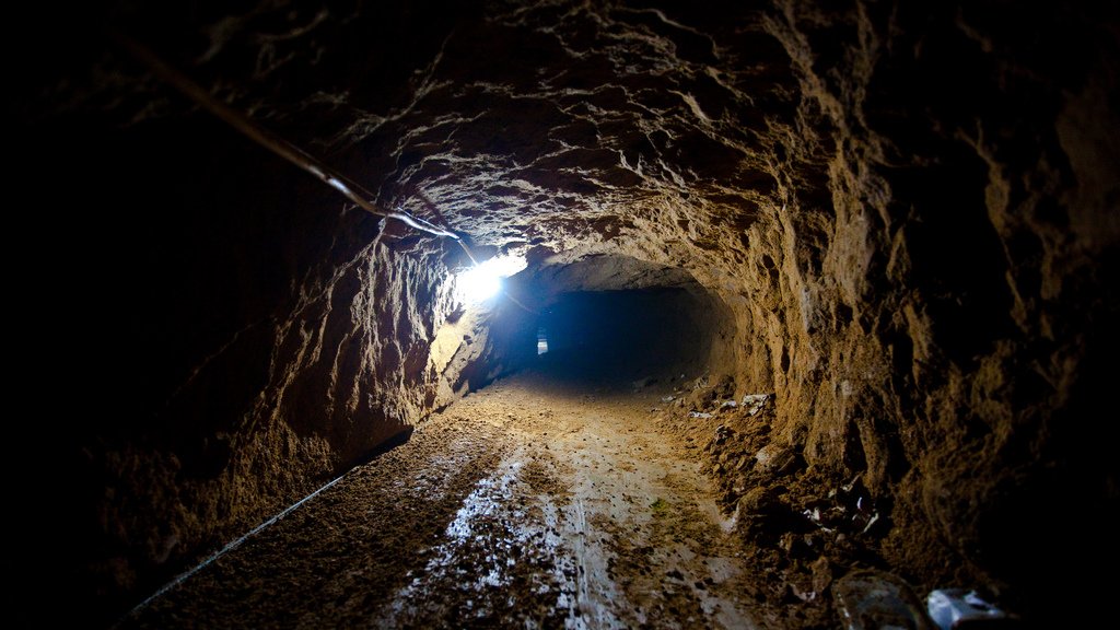 Cette image montre un tunnel souterrain. Les parois sont irrégulières et terreuses, indiquant qu'il a été creusé à la main ou à l'aide d'outils simples. Le sol est recouvert de terre, et une source de lumière artificielle éclaire l'intérieur, créant un contraste entre l'obscurité du tunnel et la lumière. À droite, on peut apercevoir des déchets éparpillés, ce qui suggère que l'endroit a été utilisé pour un certain temps. Le tunnel semble relativement étroit et évoque une atmosphère de mystère ou d'aventure.