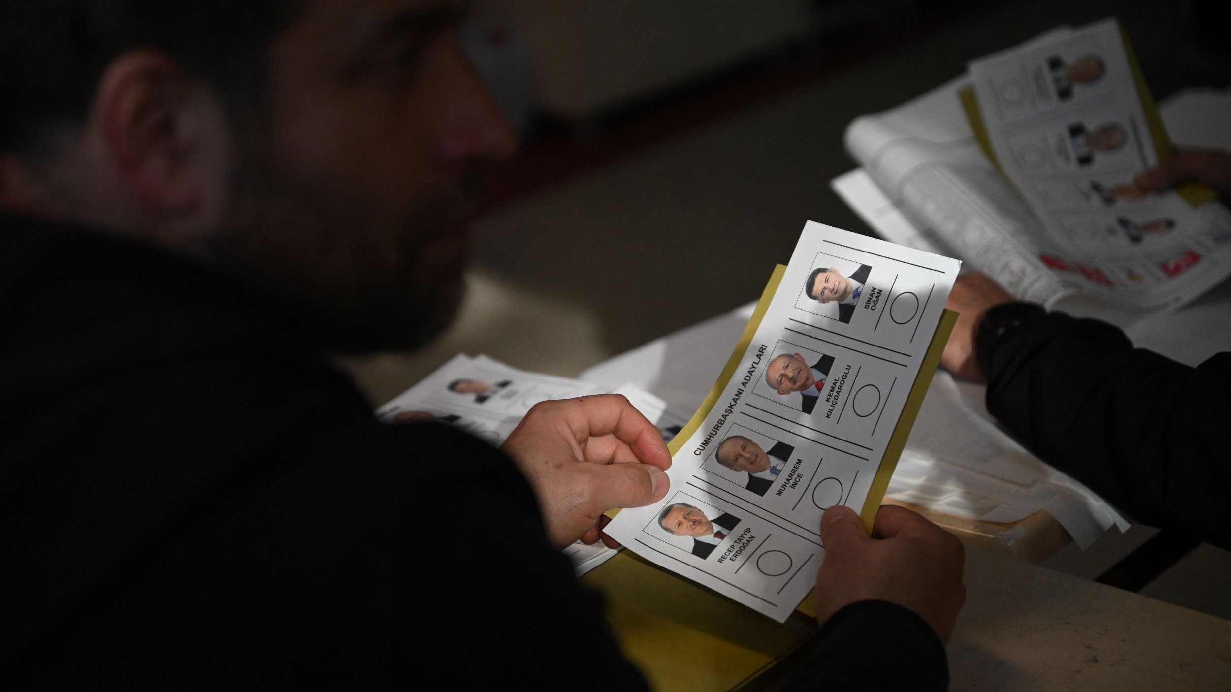The image shows a person, partially visible, holding a ballot or voting card that features photographs and names of candidates. The setting appears to be a polling station, with multiple ballots or documents visible. The individual seems to be examining the options before making a decision. The lighting suggests an indoor environment, likely during a voting process.