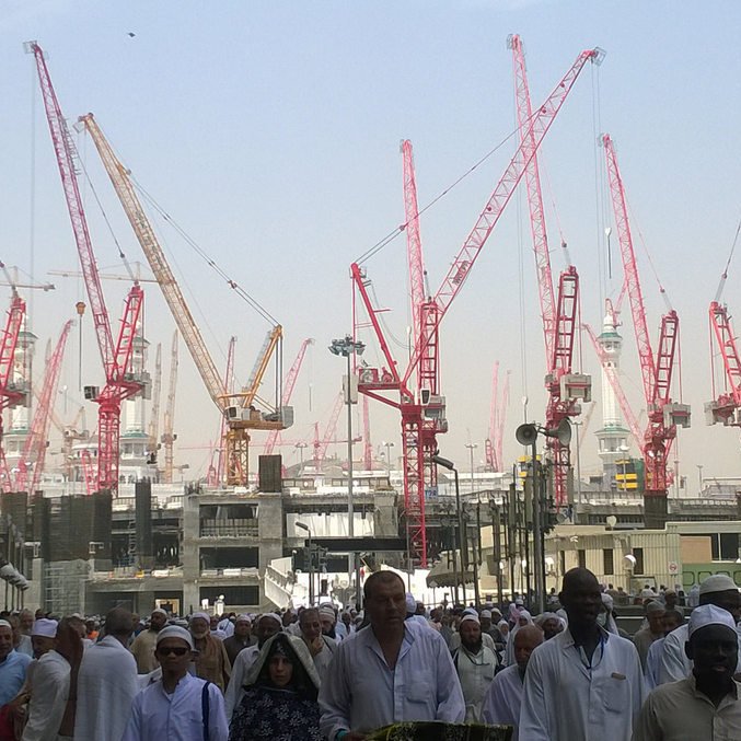 L'image montre un grand nombre de personnes, probablement des pèlerins, se déplaçant dans une zone de construction. En arrière-plan, on peut voir de nombreuses grues rouges qui soulignent les travaux en cours. Les pèlerins portent des vêtements traditionnels, souvent blancs, ce qui est courant lors des pèlerinages. L'environnement semble animé, avec une ambiance de mouvement et d'activité en raison des constructions.