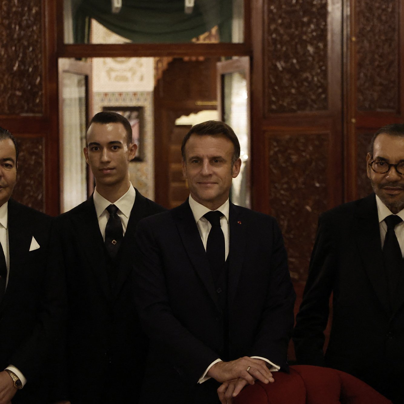 در تصویر، چهار نفر دیده می‌شوند که لباس‌های رسمی و مشکی پوشیده‌اند. در میان آن‌ها، یک مرد با کت و شلوار تیره و کراوات، در مرکز تصویر قرار دارد. او ظاهرا به عنوان یک شخصیت مهم شناخته می‌شود. دو نفر دیگر در سمت چپ و راست او ایستاده‌اند و لباس‌های مشابهی دارند. همچنین یک نفر جوان‌تر در کنار آن‌ها قرار دارد. محیط تصویر به نظر می‌رسد در یک مکان رسمی یا لوکس باشد و دکوراسیون و جزئیات آن توجه‌برانگیز است. نورپردازی نرم و گرمی دارد.