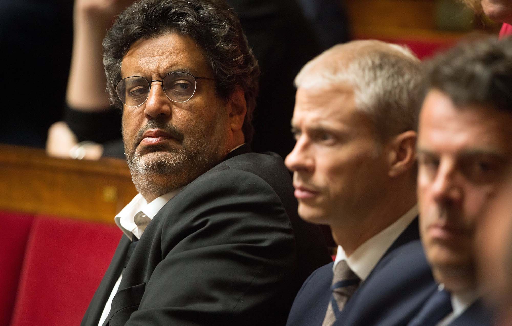 L'image montre un groupe d'hommes assis dans un cadre formel, probablement lors d'une séance législative. Un homme au premier plan porte des lunettes et un costume sombre, affichant une expression sérieuse, tandis que les autres à ses côtés semblent également attentifs et concentrés. L'arrière-plan montre des personnes floues, suggérant une ambiance politique. Les sièges sont ornés de rouge, typiques des assemblées.