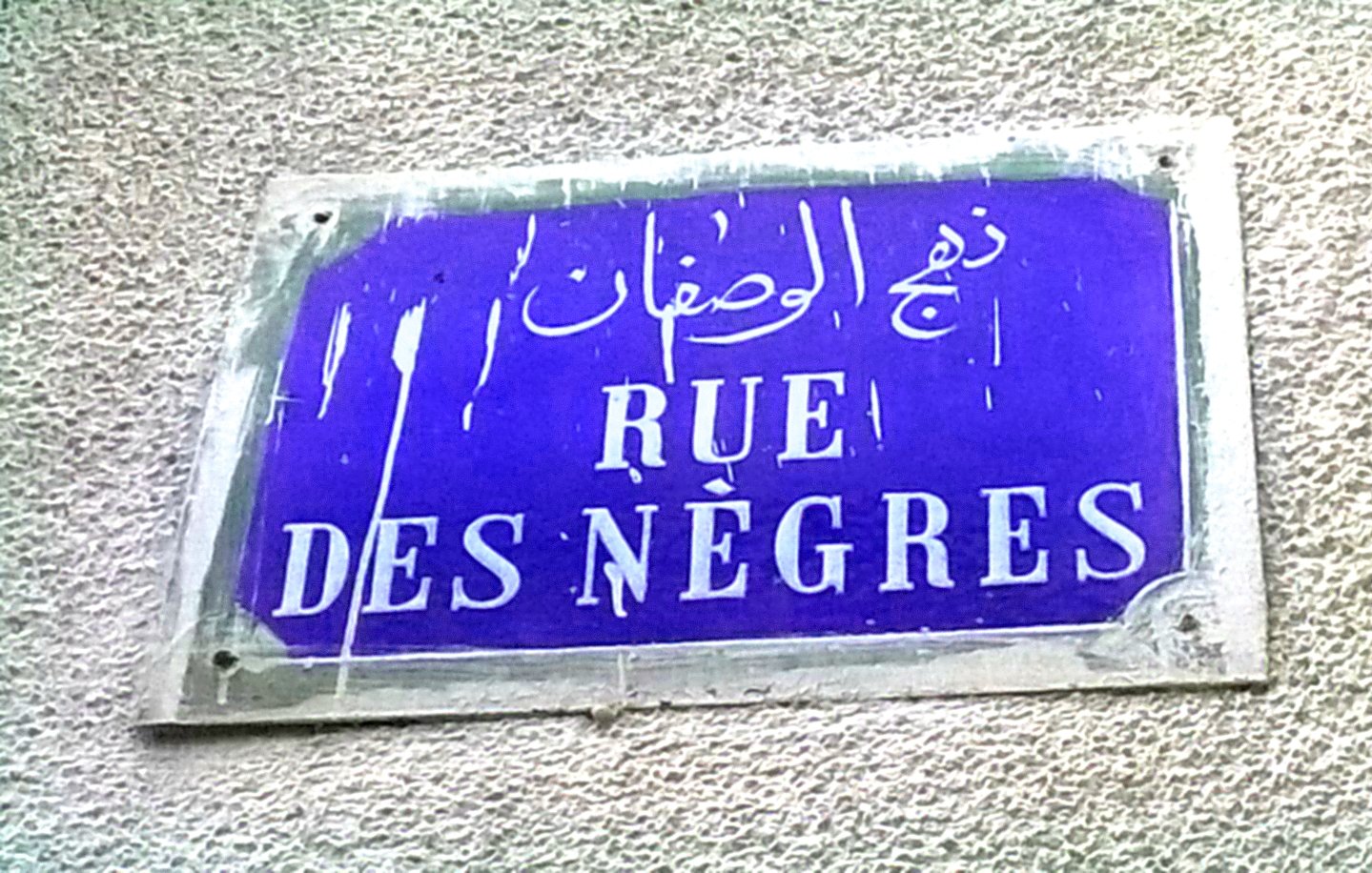 تصویر یک تابلو خیابان را نمایش می‌دهد که دارای نوشته‌هایی به زبان‌های عربی و فرانسوی است. زمینه تابلو به رنگ آبی است و نوشته‌ها به رنگ سفید. در قسمت بالای تابلو، متن عربی وجود دارد که در پایین آن، نام خیابان به زبان فرانسوی (Rue des Nègres) نوشته شده است. تابلو به نظر می‌رسد که قدیمی است و ممکن است در معرض عوامل جوی قرار داشته باشد، زیرا آثاری از کثیفی یا ساییدگی بر روی آن مشاهده می‌شود.