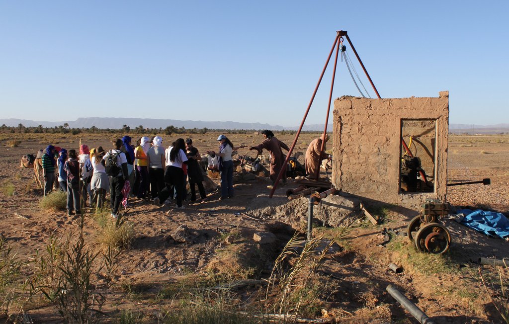 تظهر الصورة مجموعة من الأشخاص يتجمعون حول هيكل قديم في منطقة مفتوحة. الخلفية توضح منظرًا طبيعياً مع بعض التضاريس البعيدة مثل الجبال. الهيكل يبدو كأنه بناء غير مكتمل أو مهدم، ويمكن أن يكون جزءًا من مشروع أو نشاط معين. يبدو أن المشاركين يتابعون أو يتعلمون شيئًا ما متعلقًا بالهيكل. الأجواء واضحة ومشرقة، مما يضفي شعورًا بالحيوية على المشهد.