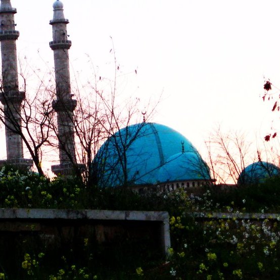 L'image montre un paysage au coucher du soleil avec deux minarets élancés se dressant vers le ciel. En arrière-plan, on peut apercevoir un grand dôme turquoise, probablement d'une mosquée, entouré de végétation. Des fleurs sauvages poussent au premier plan, ajoutant une touche de couleur et de vie à la scène. Les teintes douces du coucher de soleil créent une atmosphère paisible et tranquille.