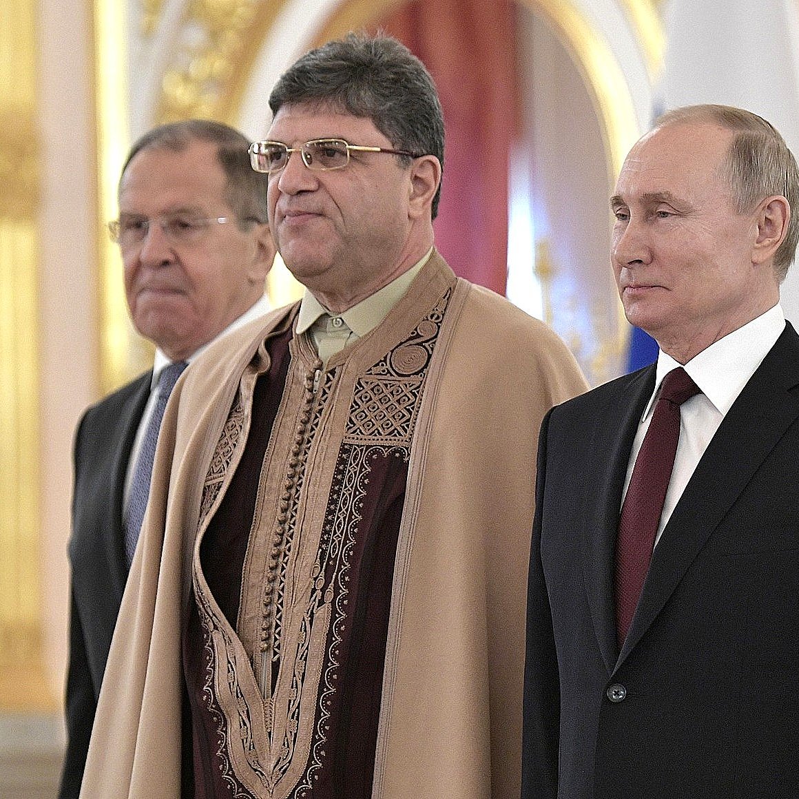 The image shows three men standing together in what appears to be an official setting, likely a ceremonial event. The man on the left is dressed in a suit, while the man in the middle is wearing traditional attire, featuring intricate patterns. The man on the right, who is likely a well-known political figure, is also dressed in a formal suit. The background is ornate, suggesting a formal and significant atmosphere.