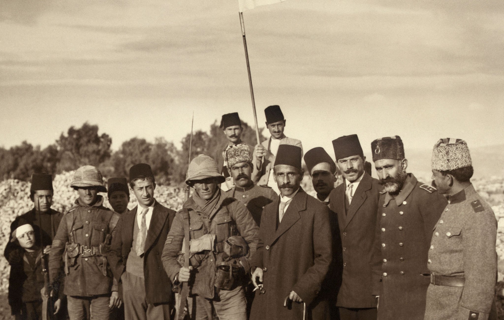 L'image montre un groupe d'hommes assemblés sur un terrain souvent aride ou montagneux. Parmi eux, certains portent des uniformes militaires, tandis que d'autres sont habillés de manière plus civile. Un homme se tient au centre, tenant un drapeau blanc, ce qui pourrait symboliser une trêve ou un cessez-le-feu. Les visages des hommes montrent une certaine gravité, tandis qu'en arrière-plan, le paysage est peuplé de collines. L'ambiance de la scène semble chargée d'une signification historique ou politique.