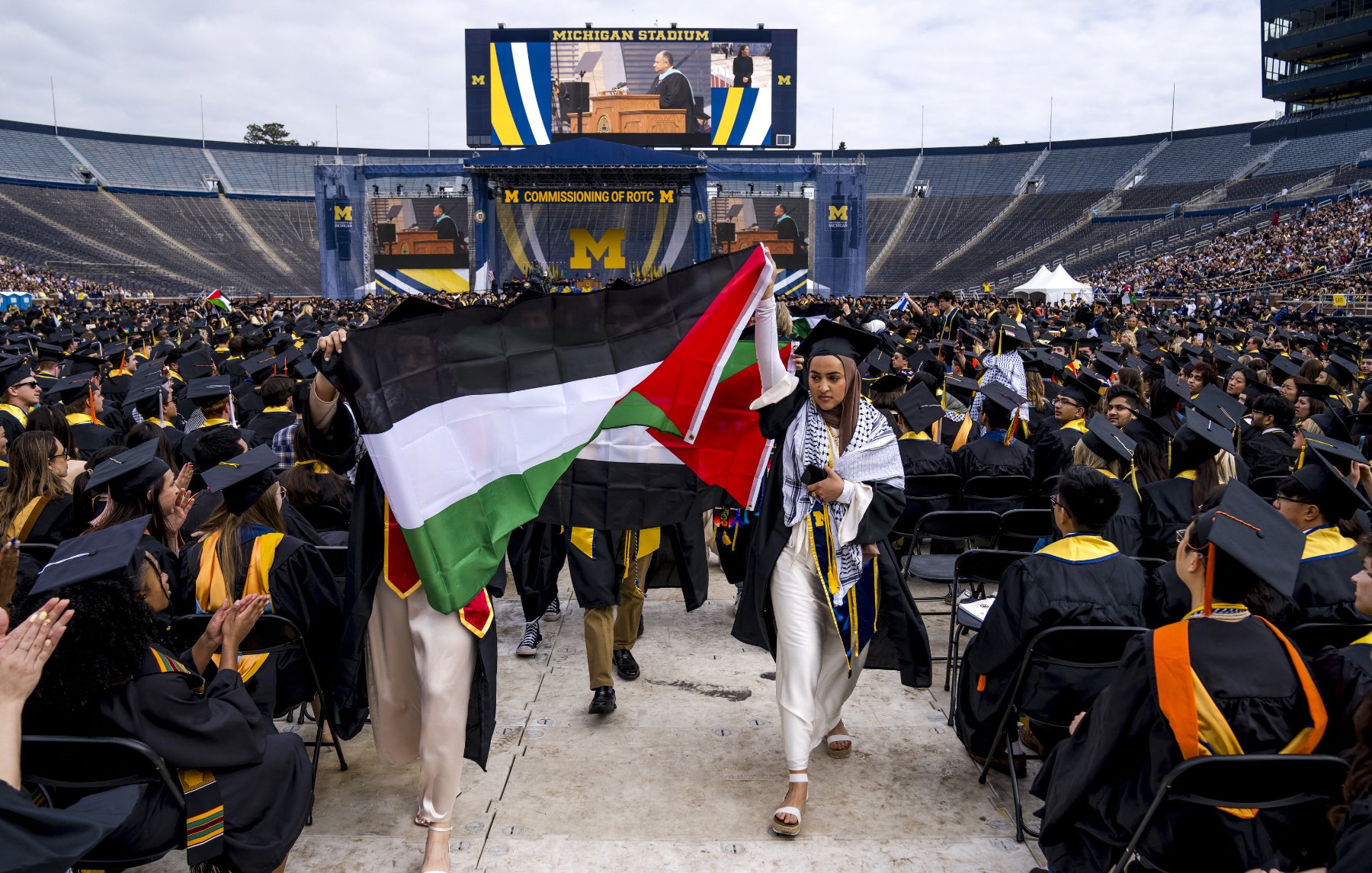 L'image montre une cérémonie de remise de diplômes qui se déroule dans un grand stade, probablement le Michigan Stadium. Des diplômés portant des toges et des chapeaux de cérémonie sont assis en rangées. Au premier plan, deux personnes tiennent des drapeaux : l'un est le drapeau palestinien et l'autre semble être un drapeau avec des couleurs distinctives. À l'arrière-plan, un grand écran affiche un orateur. L'ambiance est festive et solennelle, typique des cérémonies de graduation.