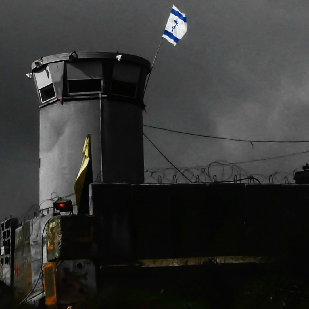 L'image montre une structure semblable à une torre de surveillance, située dans un environnement sombre et nuageux. On peut voir un drapeau israélien flottant au sommet de la tour. Des barbelés sont visibles autour du périmètre, ce qui suggère une zone sécurisée ou protégée. L'éclairage de la scène crée une atmosphère de tension, accentuant l'aspect austère du lieu.