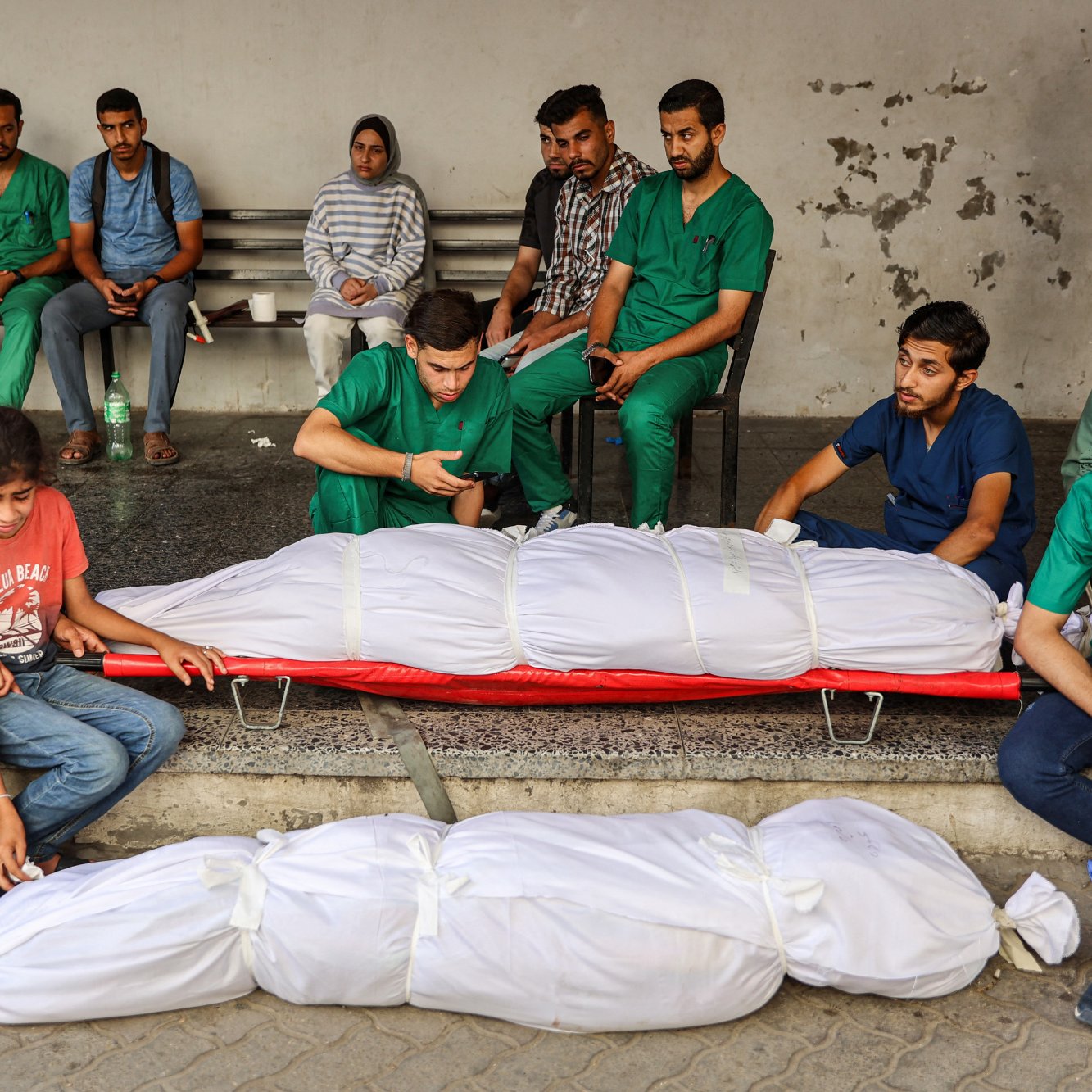 L'image montre un groupe de personnes assises sur des bancs, visiblement en attente dans un espace public. Parmi elles, certains portent des vêtements de santé, tandis que d'autres sont habillés de manière quotidienne. Au premier plan, deux corps enveloppés dans des draps blancs sont visibles, accompagnés de personnes qui semblent éprouver de la tristesse. L'atmosphère générale dégage une forte émotion, suggérant un moment de deuil ou de perte.