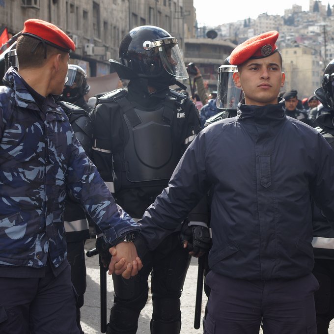 L'image montre un groupe de policiers ou de soldats, avec des uniformes distinctifs, se tenant les mains. Au centre, un homme en majorette avec un béret se distingue. Derrière eux, des membres des forces de l'ordre portent des casques et des équipements de protection. L'environnement semble urbain, avec des bâtiments en arrière-plan. L'atmosphère peut suggérer une situation de tension ou de manifestation.