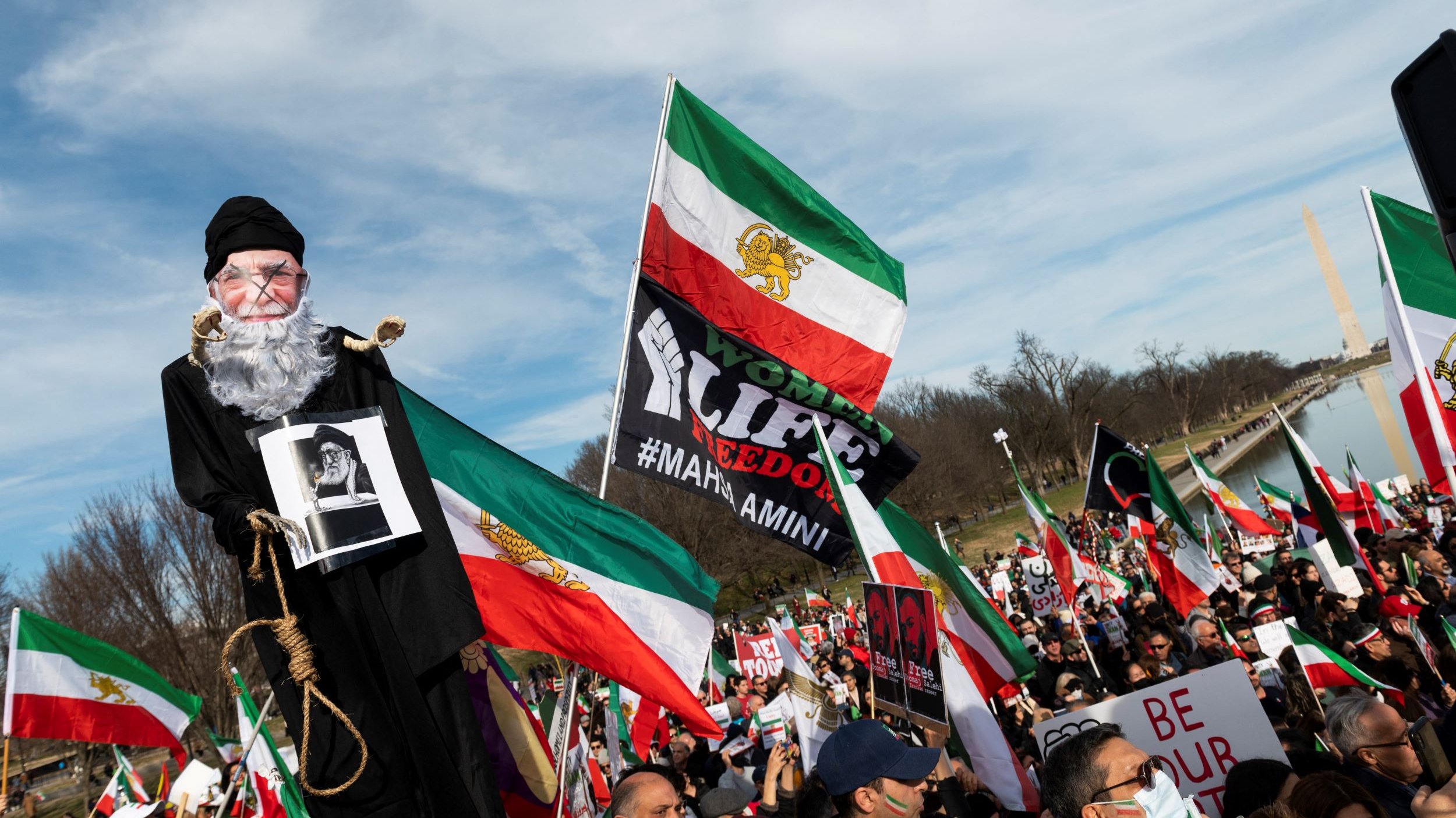 L'immagine ritrae una manifestazione in cui si vedono molte persone riunite, molte delle quali tengono in mano bandiere dell'Iran. C'è un grande cartello con la scritta "Woman, Life, Freedom" e un uomo che tiene una sagoma con la figura di un anziano, che sembra simboleggiare una figura autoritaria. Sullo sfondo si possono notare manifestanti con vari cartelli e bandiere, tutti uniti per esprimere il loro messaggio di protesta. La scena trasmette un forte senso di unità e determinazione tra i partecipanti.