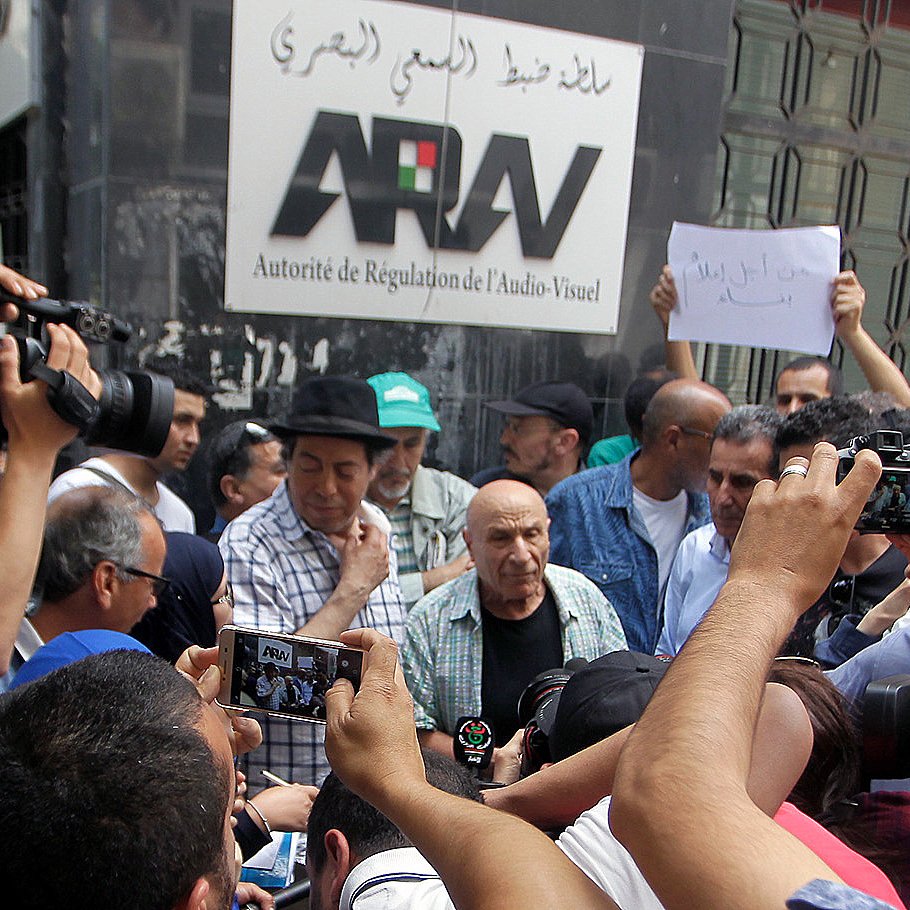L'image montre un groupe de personnes rassemblées autour d'un homme au premier plan, visiblement en pleine discussion ou confrontation. Plusieurs caméras et téléphones portables sont levés, capturant l'événement. En arrière-plan, on peut voir une bannière avec le mot "ARAV" qui fait référence à l'Autorité de Régulation de l'Audiovisuel. La scène semble être un moment de protestation ou de manifestation, avec des expressions variées sur les visages des participants.