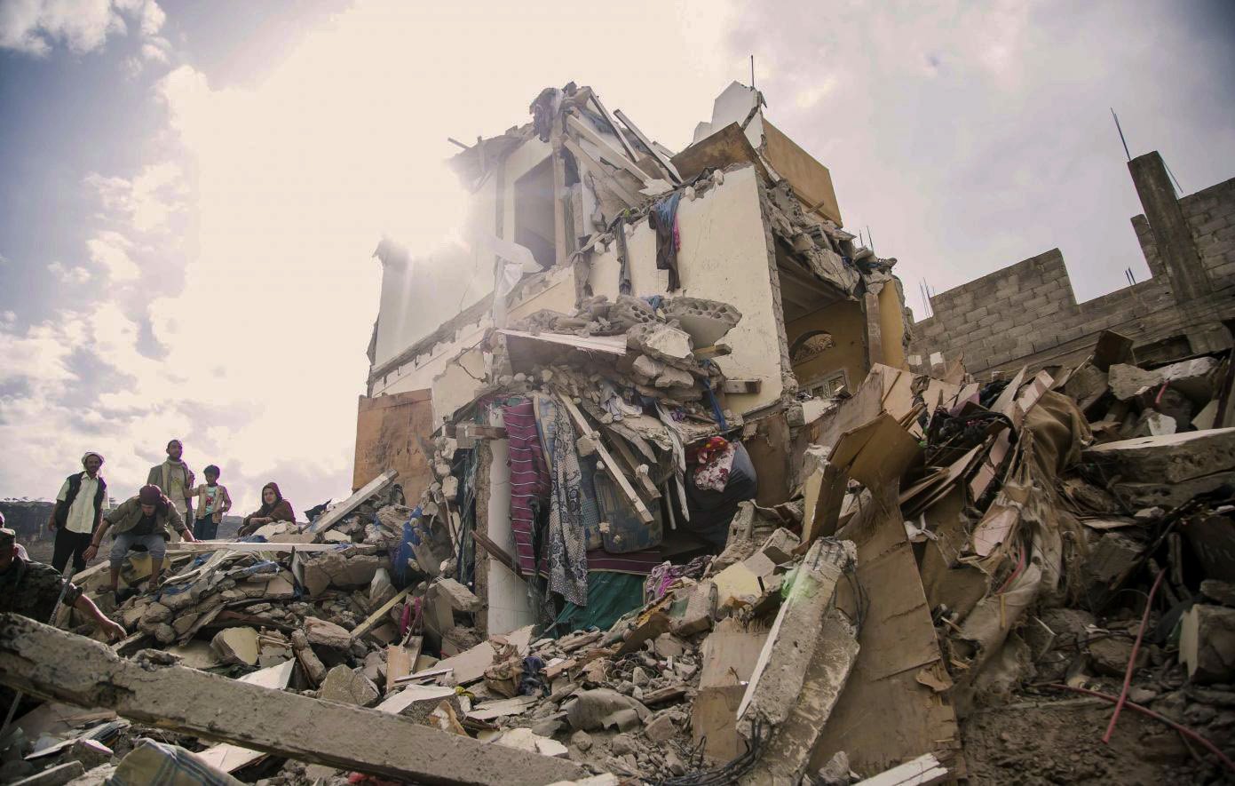 The image depicts a collapsed building, showing significant structural damage with debris scattered around. The upper portions of the structure are crumbled, and various materials like concrete, wood, and personal belongings are visible. In the foreground, there are a few individuals, likely responders or onlookers, inspecting the site. The scene is illuminated by sunlight breaking through the clouds, which adds a stark contrast to the devastation shown. The overall mood conveys a sense of disaster and the aftermath of a catastrophic event.