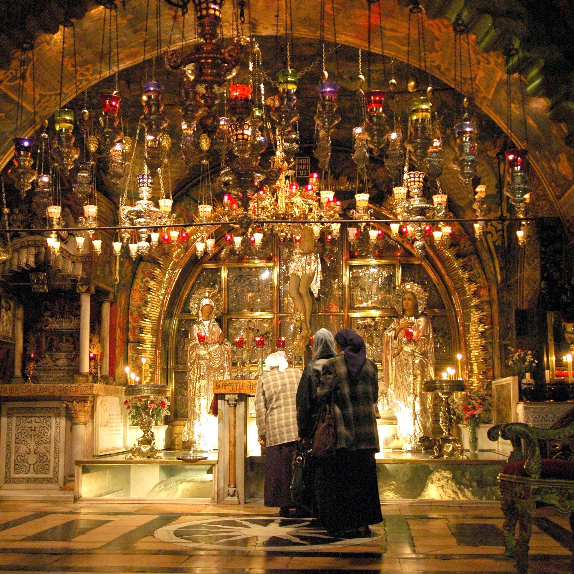 L'image montre l'intérieur d'un lieu de culte, probablement une église ou une chapelle. L'architecture est ornée, avec un plafond voûté richement décoré. On peut voir des lumières colorées suspendues et des candélabres, créant une ambiance chaleureuse et mystique. Au centre, il y a des figures dorées, possiblement des icônes ou des statues, entourées de bougies et de fleurs. Quelques personnes, vêtues de manteaux et de foulards, se tiennent devant cette scène, semblant s'engager dans une prière ou une contemplation.
