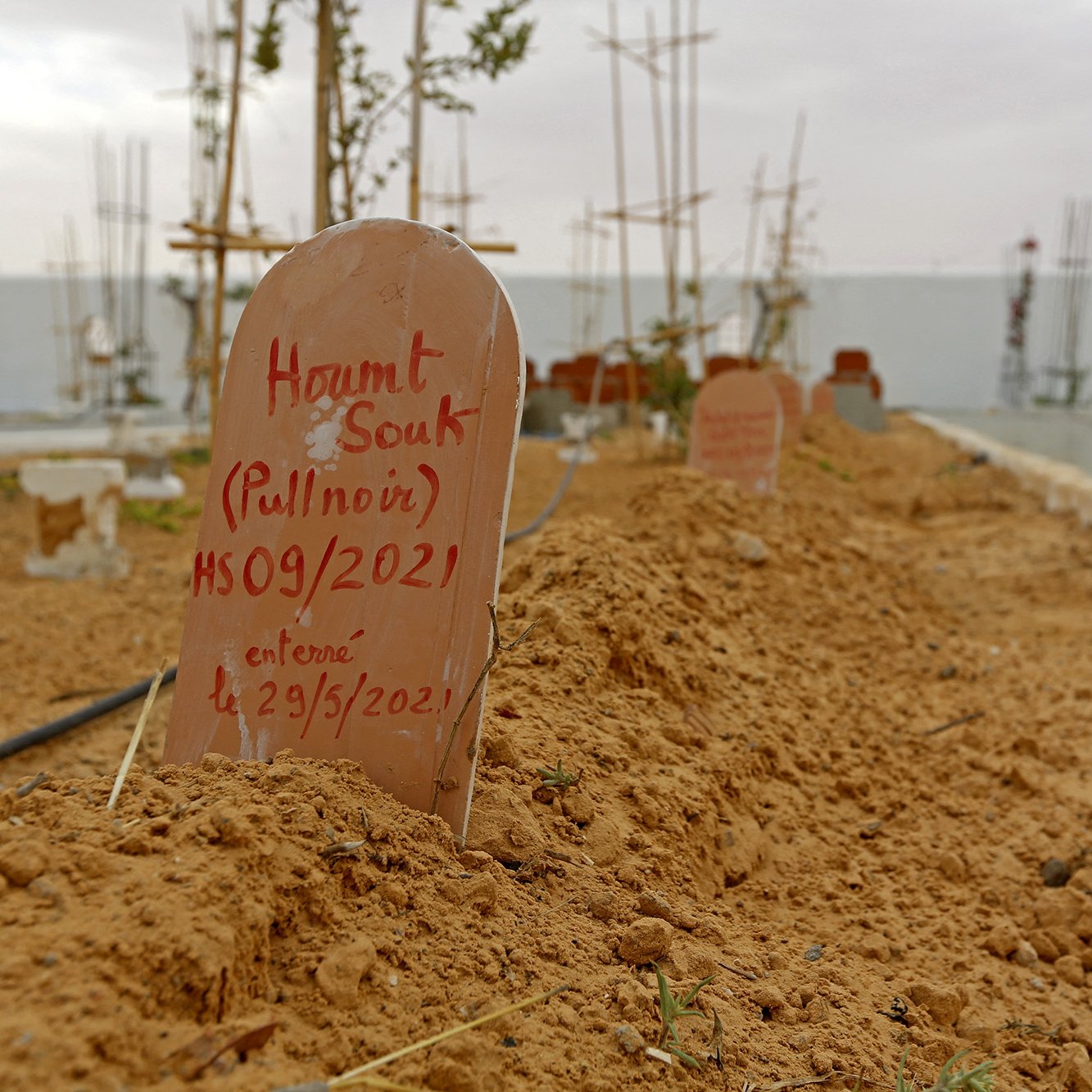 L'image montre un cimetière avec des tombes marquées par des plaques en terre cuite. Le sol est sablonneux et légèrement irrégulier, avec quelques plantes éparses. En arrière-plan, on remarque des structures en construction, probablement des bâtiments ou des monuments. Le ciel semble nuageux, créant une atmosphère calme mais mélancolique. Les inscriptions sur les plaques indiquent les noms et des dates, suggérant que ces êtres chers ont été récemment enterrés.