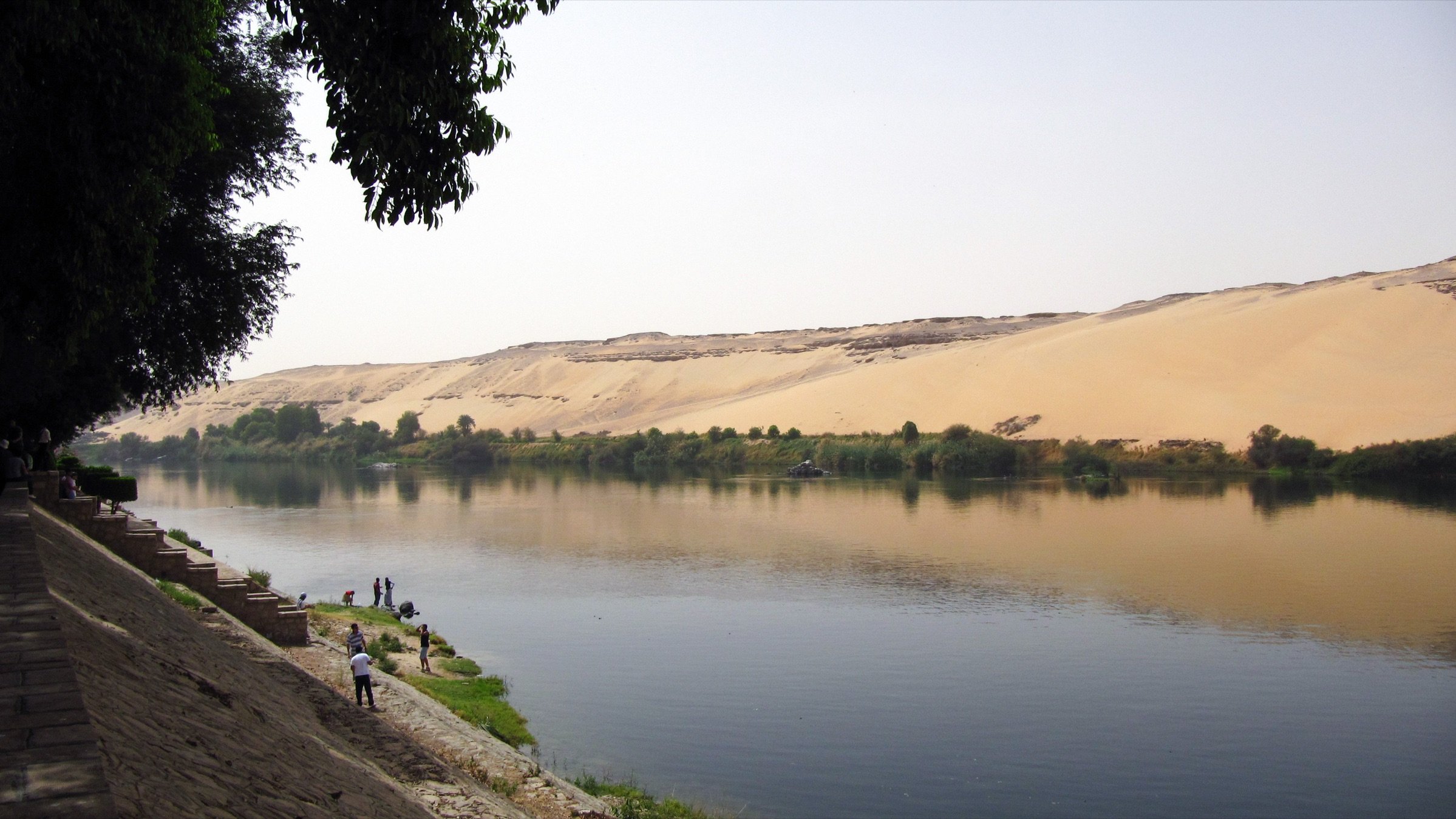 L'image montre un paysage paisible où un fleuve serpente le long d'une rive. Sur un côté, il y a des dunes de sable doré qui montent en pente, créant un contraste avec la verdure des arbres qui bordent le fleuve. Des personnes se trouvent au bord de l'eau, peut-être en train de pêcher ou de profiter de la nature. Le ciel est clair, et la lumière dubaine donne une atmosphère sereine à la scène.