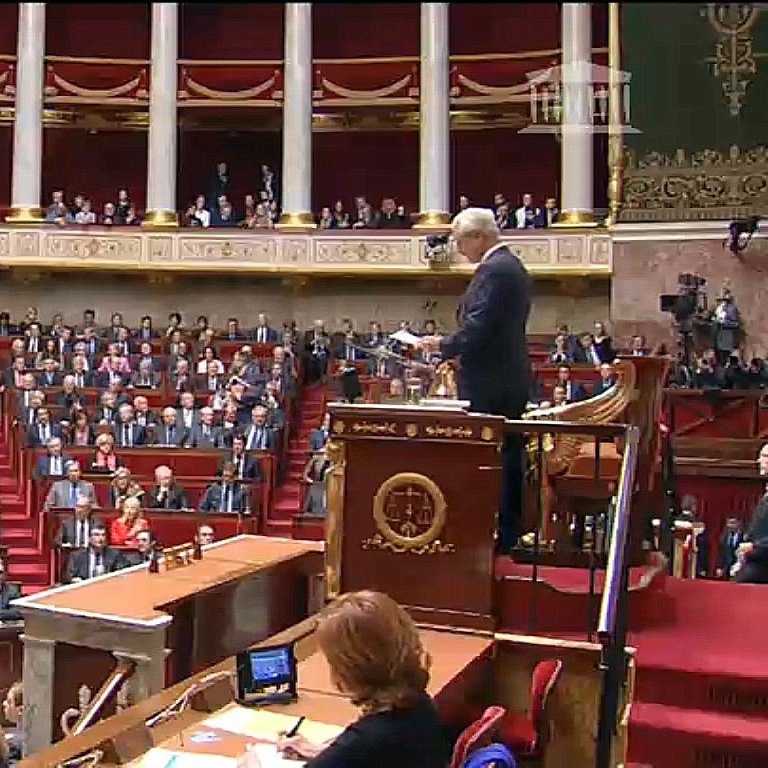L'image montre une scène à l'Assemblée nationale, un lieu de débat politique en France. On y voit un grand amphithéâtre avec de nombreux sièges, occupés par des députés. Au premier plan, un homme se tient debout devant un pupitre, probablement en train de faire un discours ou de présenter une proposition. Les députés semblent attentifs et sont disposés en plusieurs rangées, tandis que certains sont assis à des tables sur le côté. L'architecture est typiquement institutionnelle, avec des éléments ornementaux et une atmosphère solennelle.